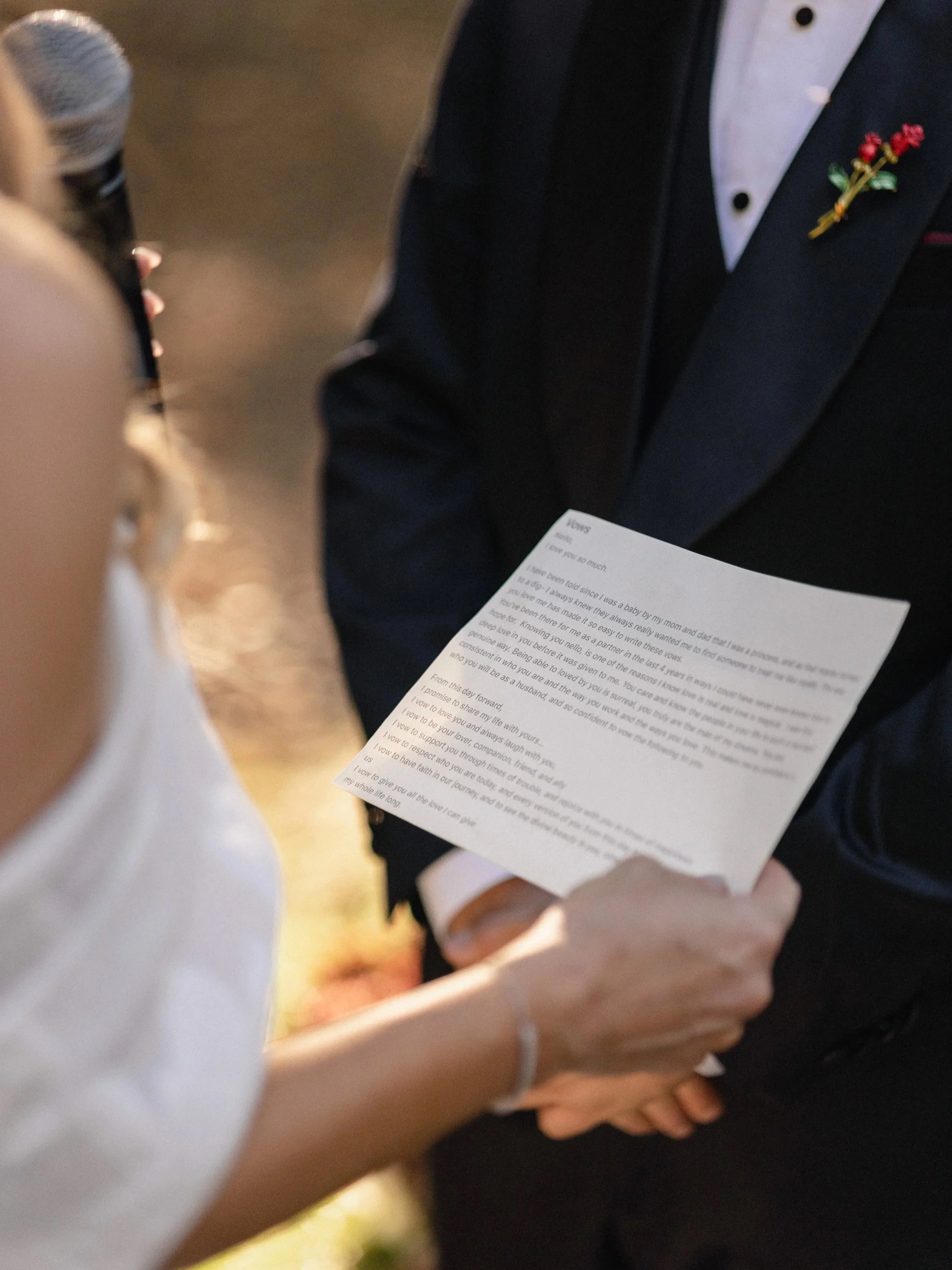A man dressed in formal attire holding a printed wedding vows note, with a boutonniere on his black suit jacket, during a wedding ceremony.