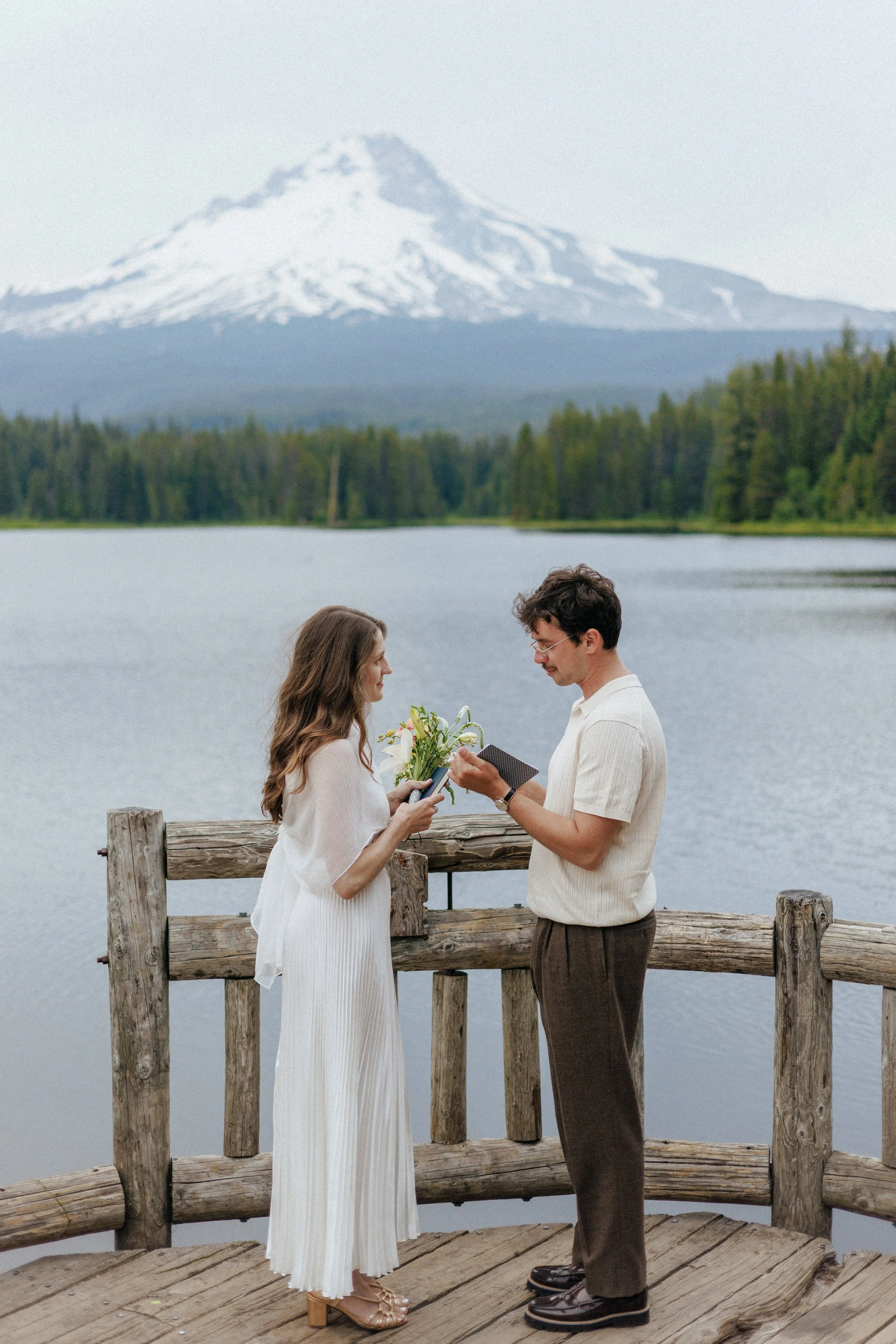 A couple stands on a wooden dock by a lake, with a mountain with snow on top in the background. The woman is holding a bouquet of flowers, and the man is holding a small book or card. They are facing each other and smiling.