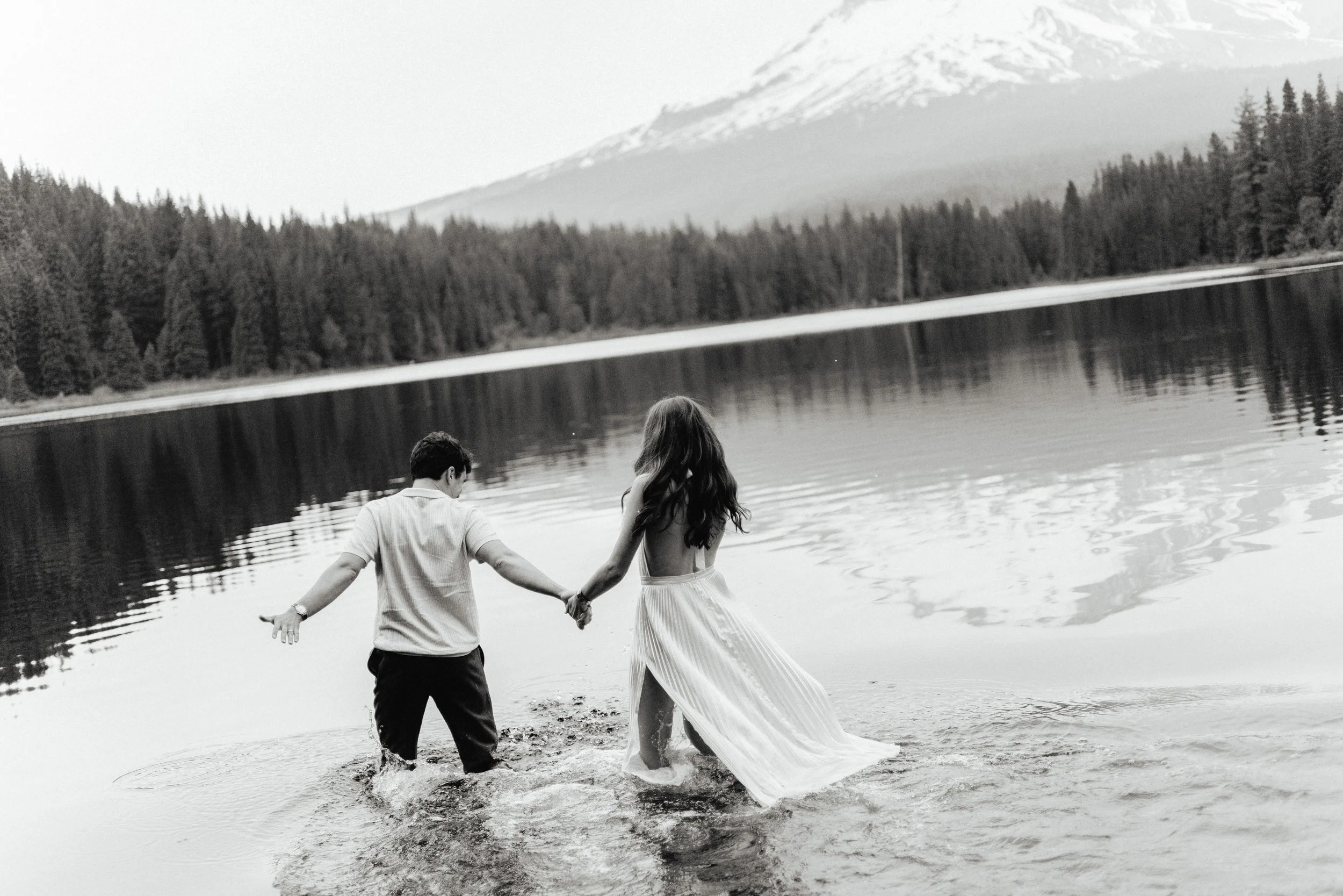 A black-and-white photo of a couple holding hands and wading into a lake, with a forested shoreline and snow-capped mountains in the background.