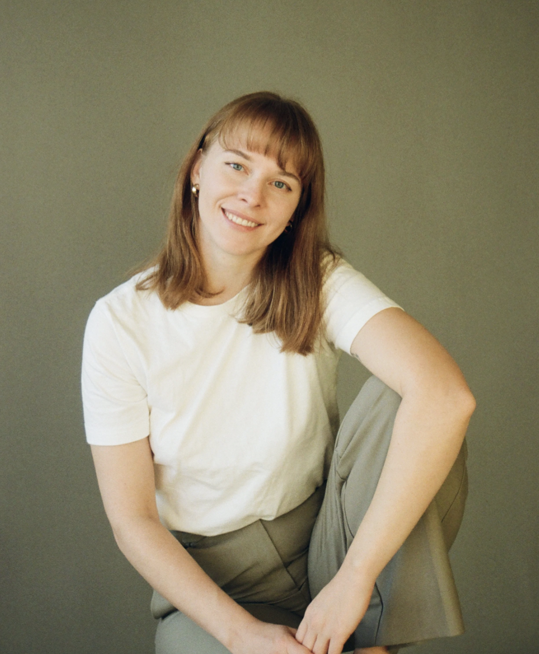 Portrait of a young woman with shoulder-length light brown hair and bangs, smiling, sitting on the floor with one knee up, wearing a white t-shirt and gray pants, against a plain neutral background.