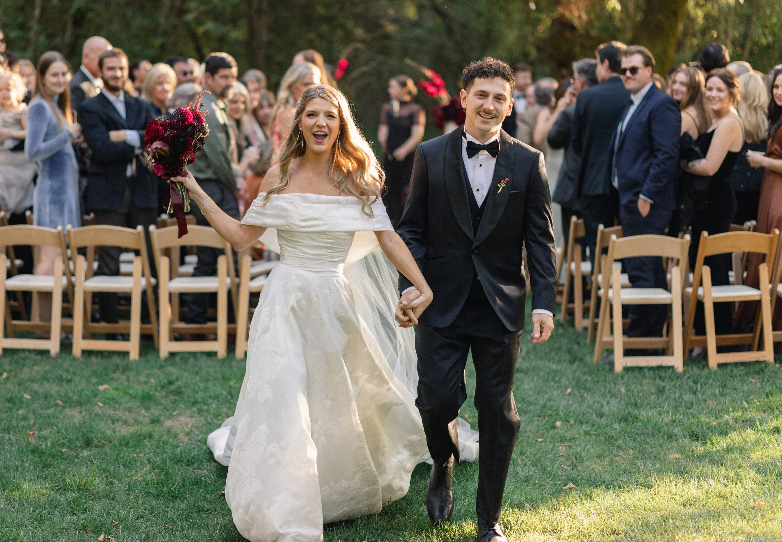A bride and groom walking hand-in-hand outdoors during a wedding ceremony, surrounded by seated and standing guests on a grassy area with trees in the background.