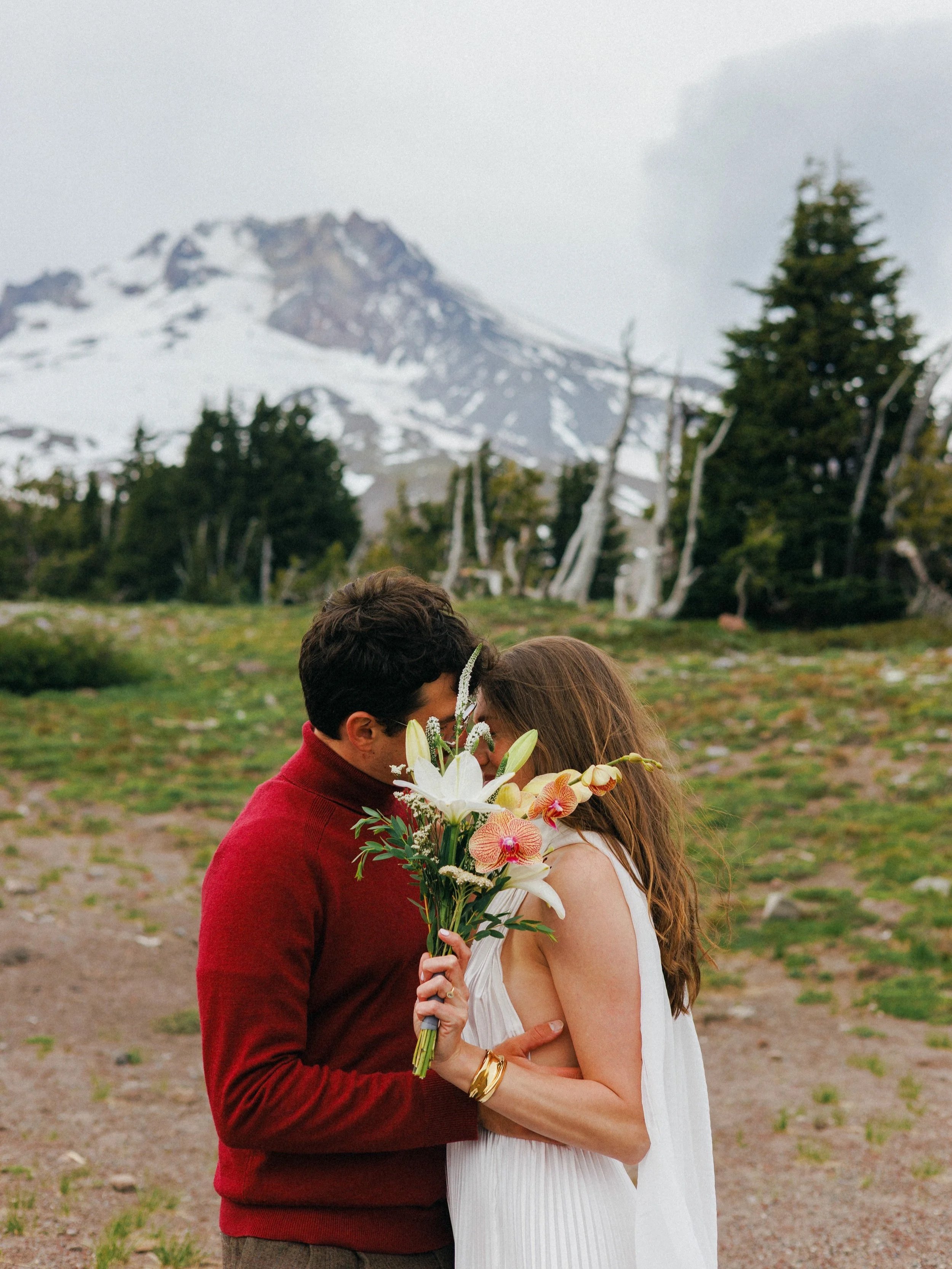 A couple sharing a kiss outdoors with a mountain and trees in the background, holding a bouquet of flowers.