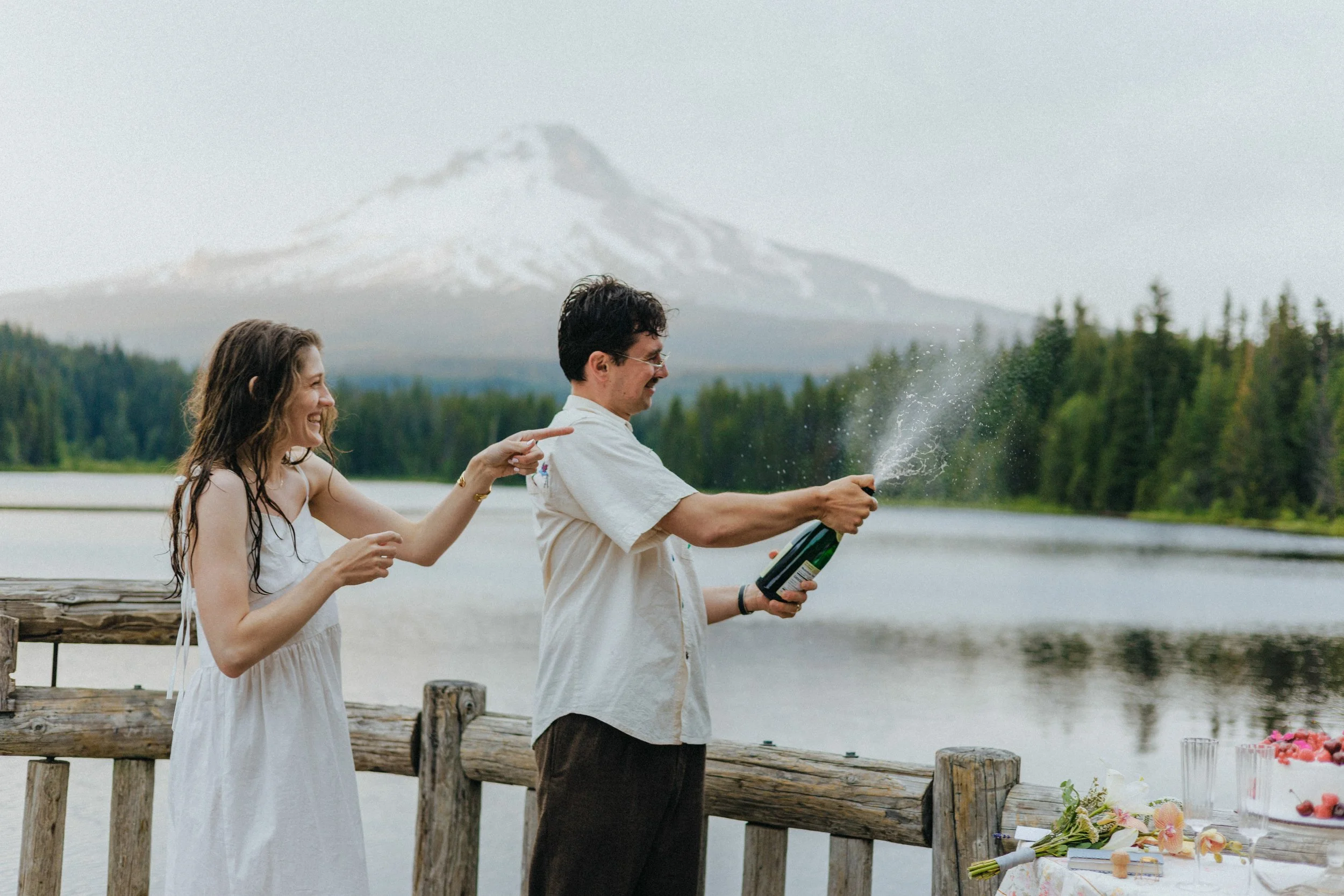 Two people are celebrating outdoors near a lake with a mountain in the background. One person is holding a champagne bottle and spraying it, while the other is smiling and pointing at it. A decorated table with flowers and a cake is visible on the right.
