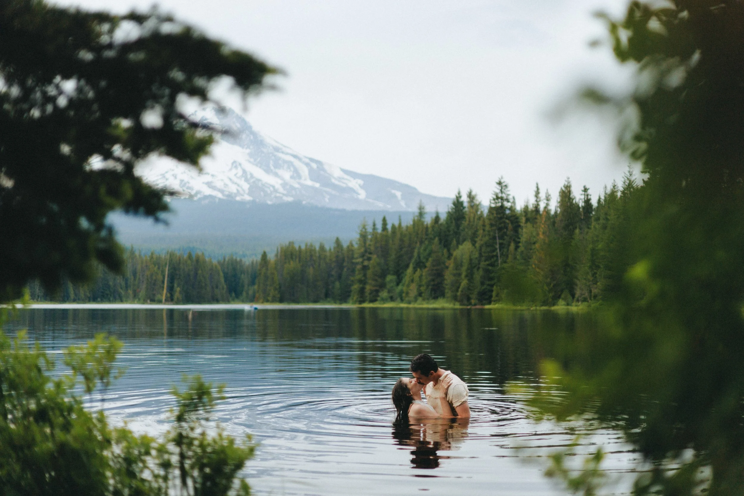A couple kissing in a lake with a forested landscape and snow-capped mountains in the background, during daytime.