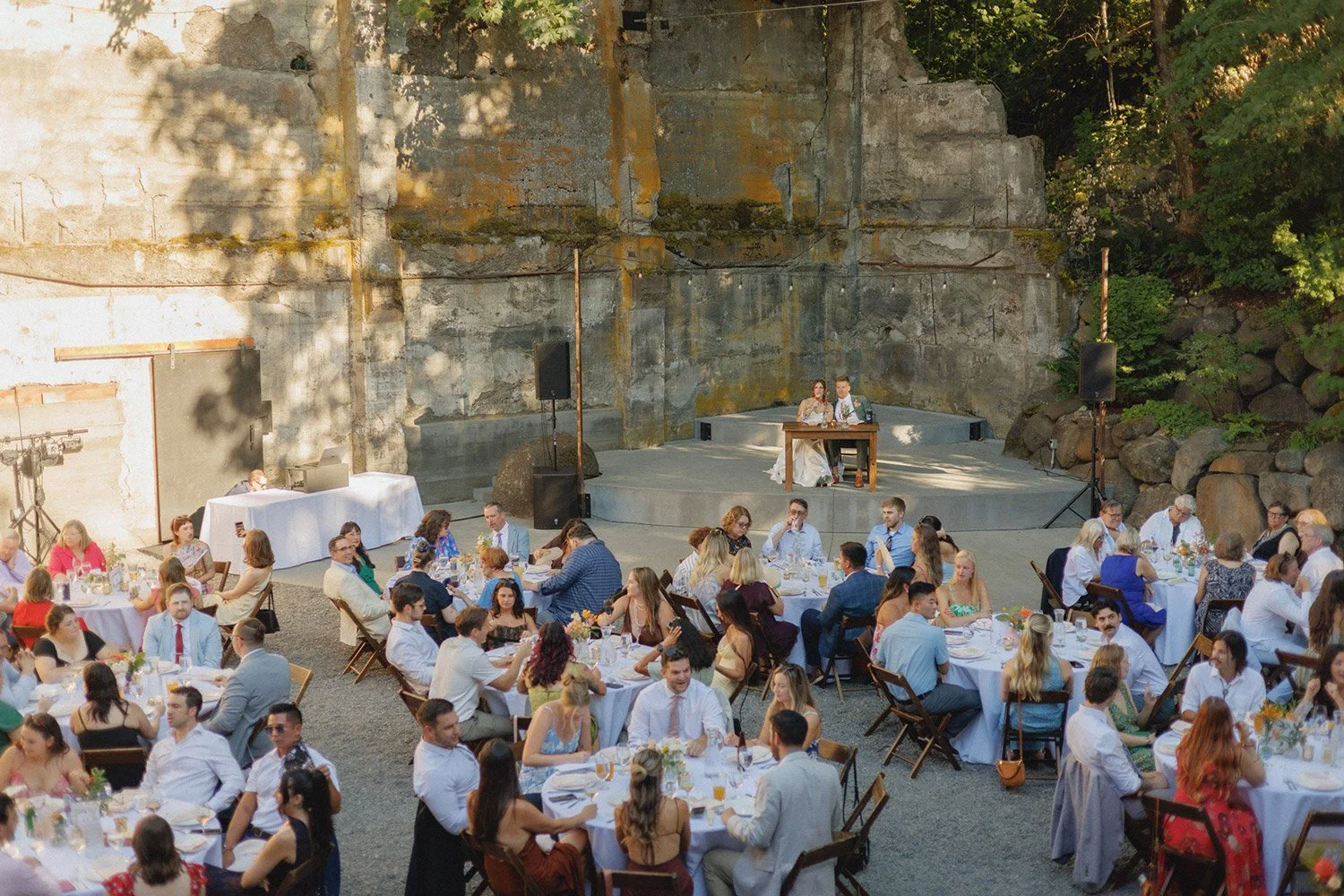 Outdoor wedding reception with guests seated at round tables covered with white tablecloths, in front of a rocky wall and surrounded by trees, with a raised stage where a bride and groom are seated.