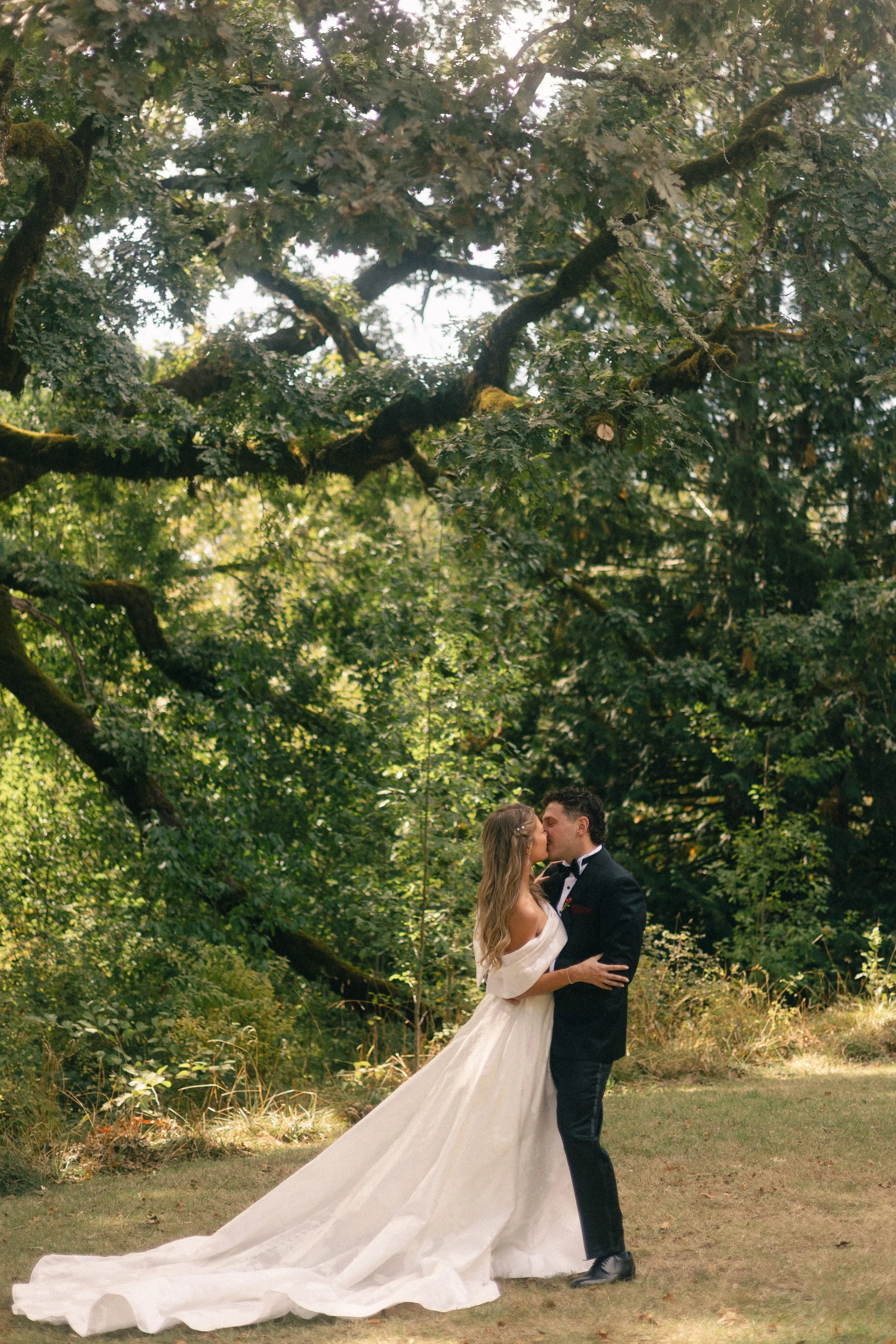 A bride and groom share a kiss outdoors in a wooded area. The bride wears a long white wedding gown with a train, and the groom wears a black tuxedo with a bow tie. They are embracing each other in a scenic, natural setting surrounded by trees and sunlight filtering through the leaves.