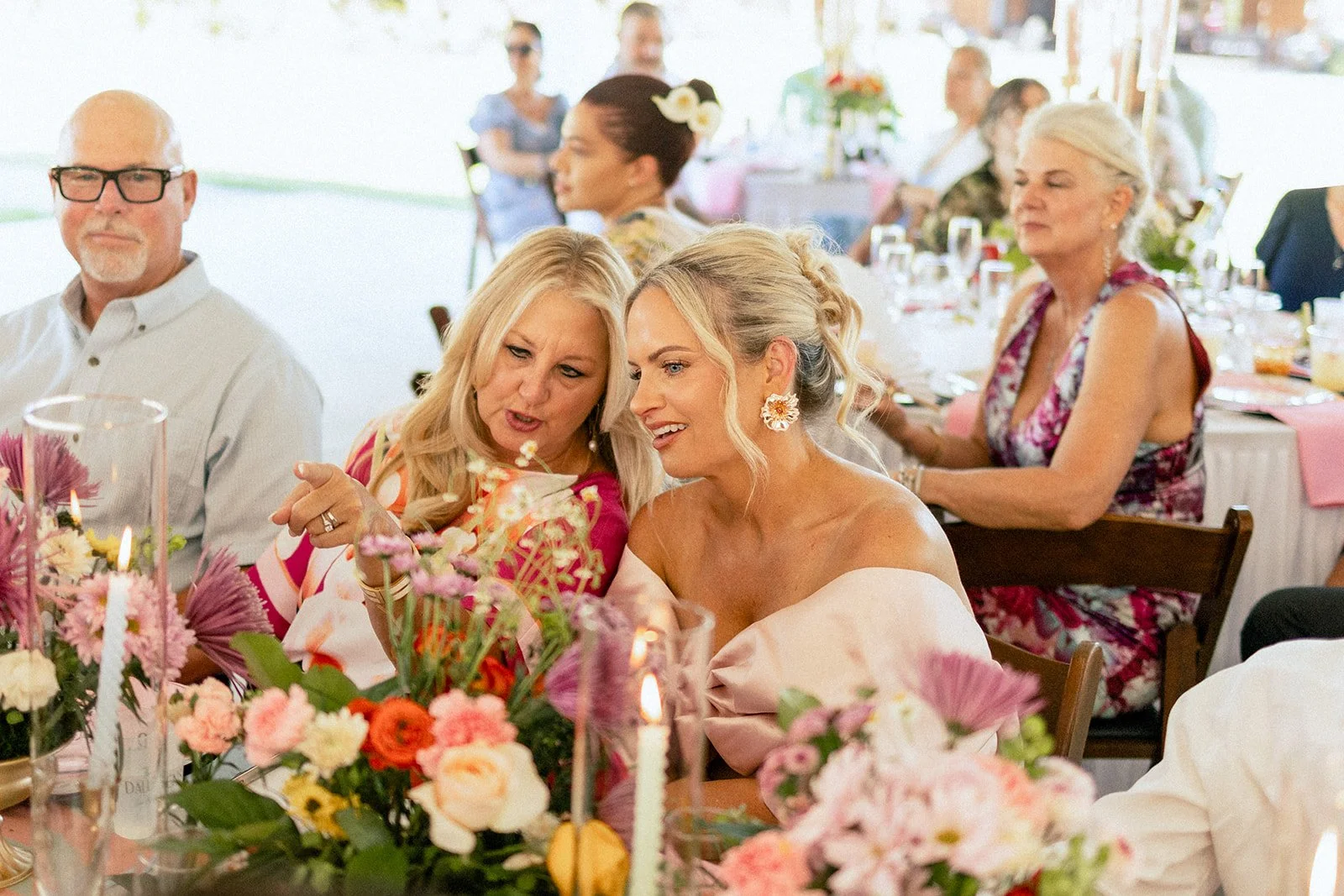 People sitting at a decorated table during a celebration, with women talking and floral arrangements in the foreground.