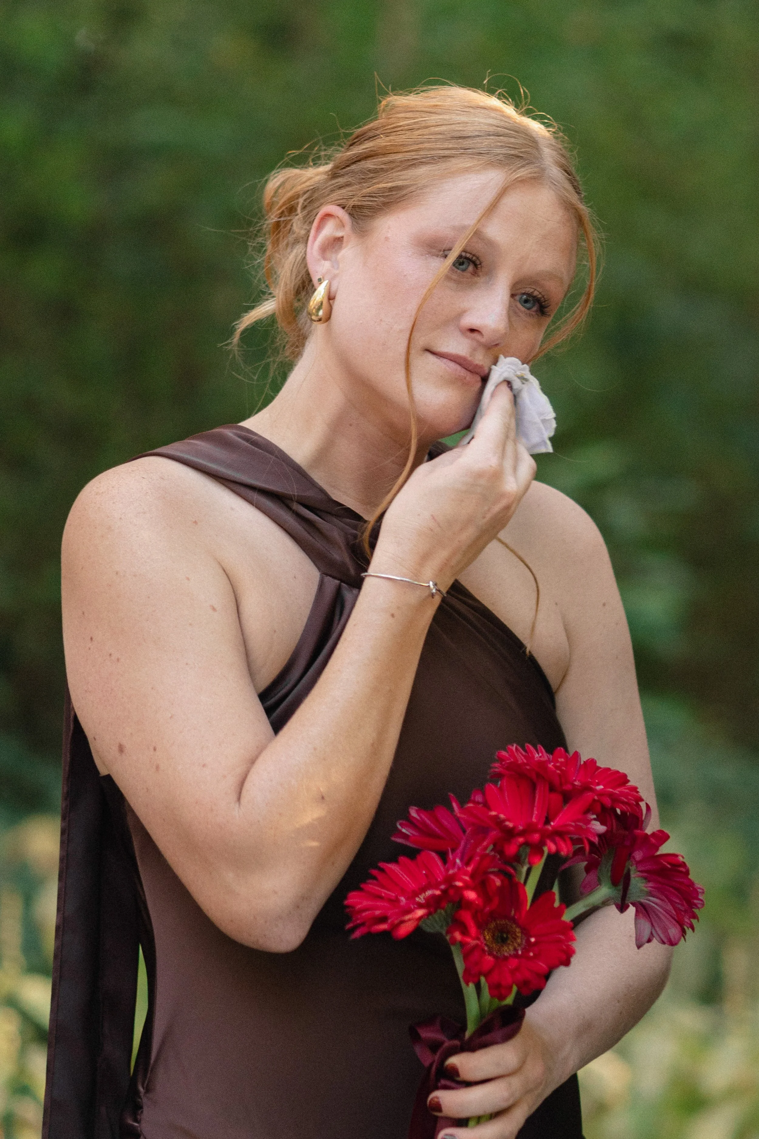 A young woman with red hair and blue eyes holding a bouquet of red and pink flowers, wearing gold hoop earrings and a sleeveless brown dress, standing outdoors with greenery in the background.