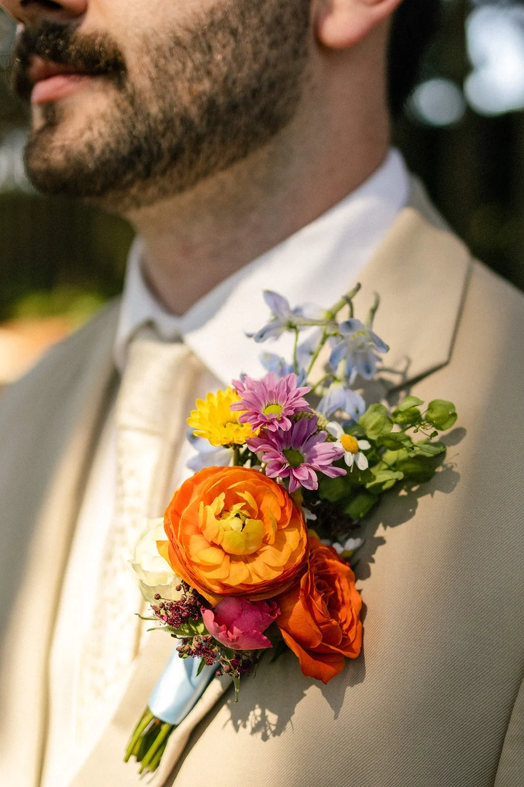 Close-up of a man's suit with a colorful boutonniere pinned on the lapel, featuring orange ranunculus, pink daisies, purple forget-me-nots, white daisies, and green foliage.