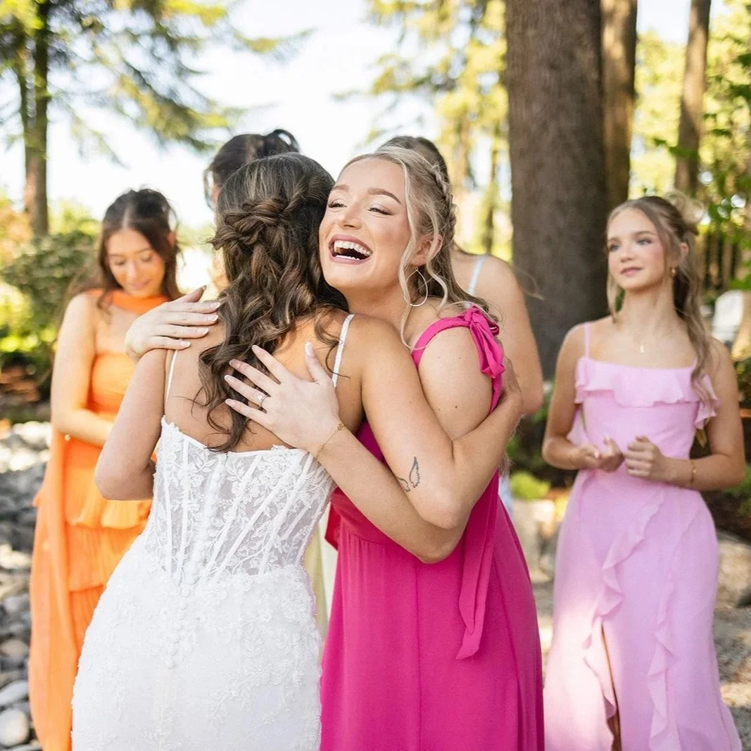 Two women hugging, one in a white dress and the other in a pink dress, smiling happily. Four other women in colorful dresses are in the background, outdoors with trees and sunlight.