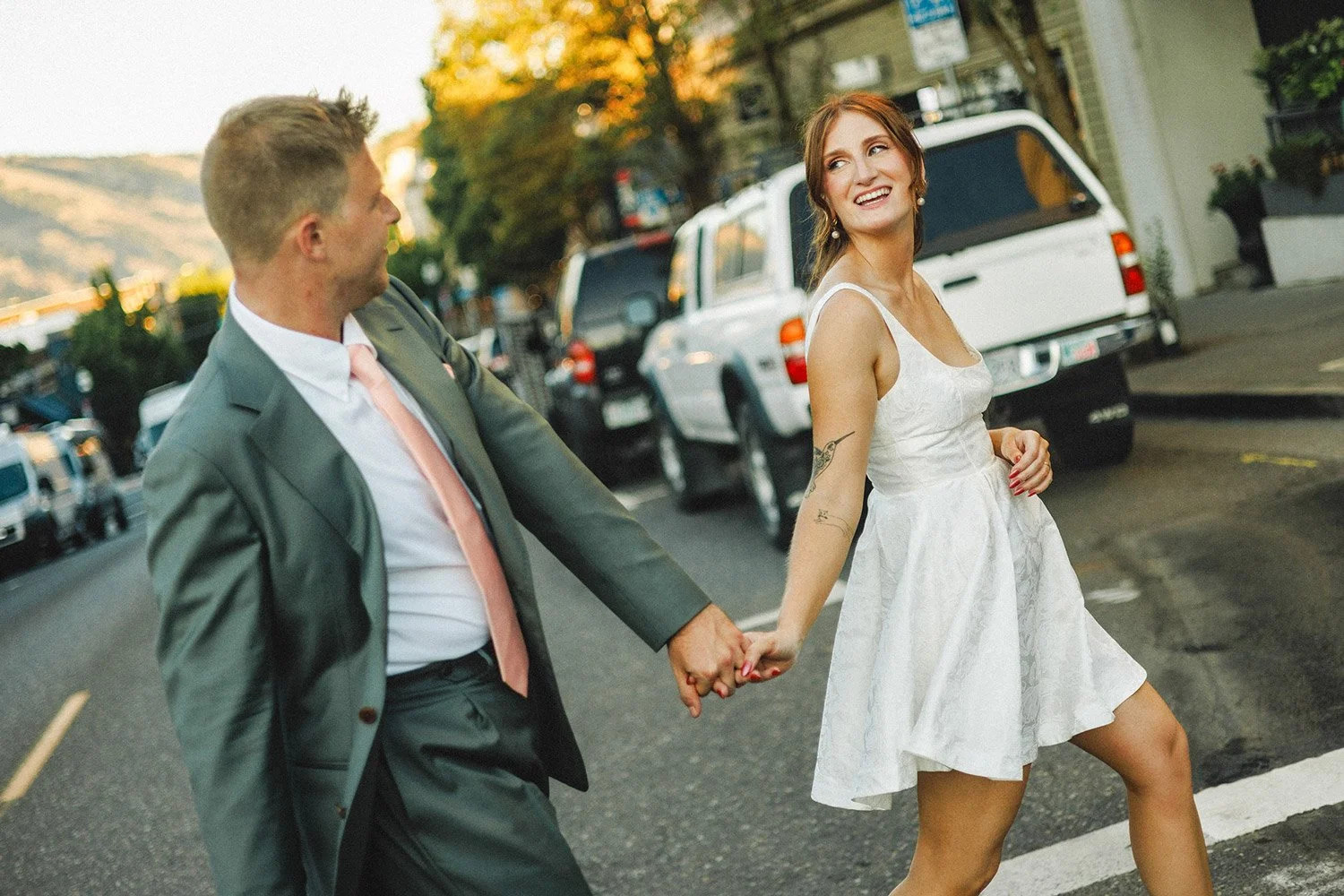A couple holding hands and walking on a city street, smiling at each other. The woman has red hair, wears a white dress, and has tattoos on her arm. The man wears a grey suit with a pink tie.