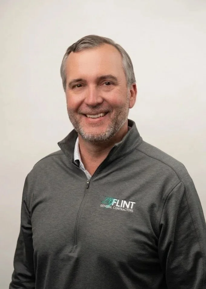 A smiling man with short gray hair and a beard wearing a dark gray button-up shirt with the logo 'LUNT GENERAL CONTRACTORS' embroidered on it, standing against a plain white background.