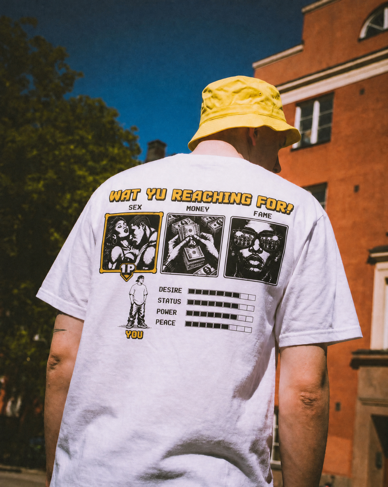 Person wearing a white t-shirt with black and yellow graphic and text, yellow bucket hat, standing outdoors near a red brick building at dusk, with trees visible in the background.