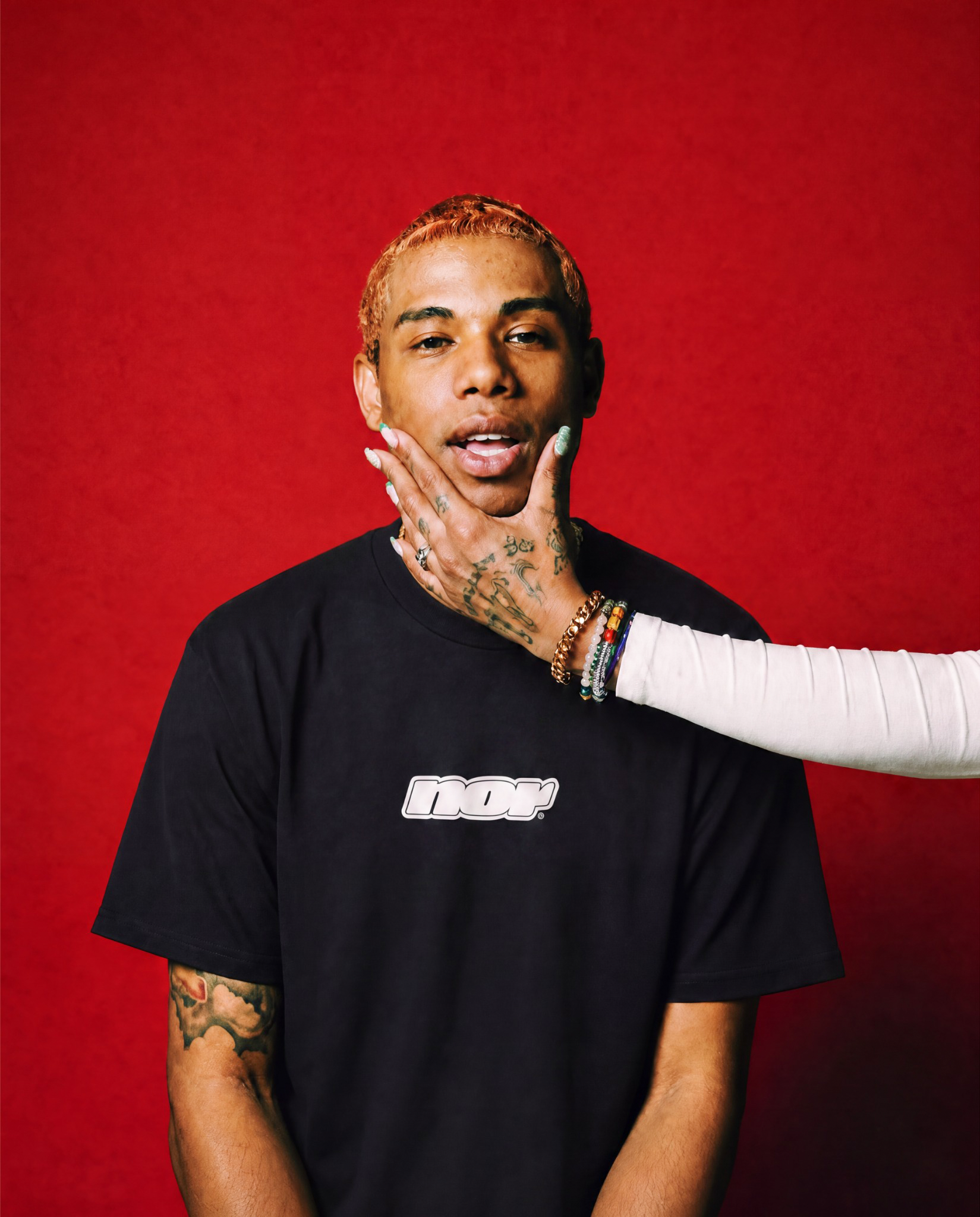 A person with tattoos on their arm wearing a black shirt with the word 'NOIR' on it. A hand on their face has colorful bracelets and manicured nails.
