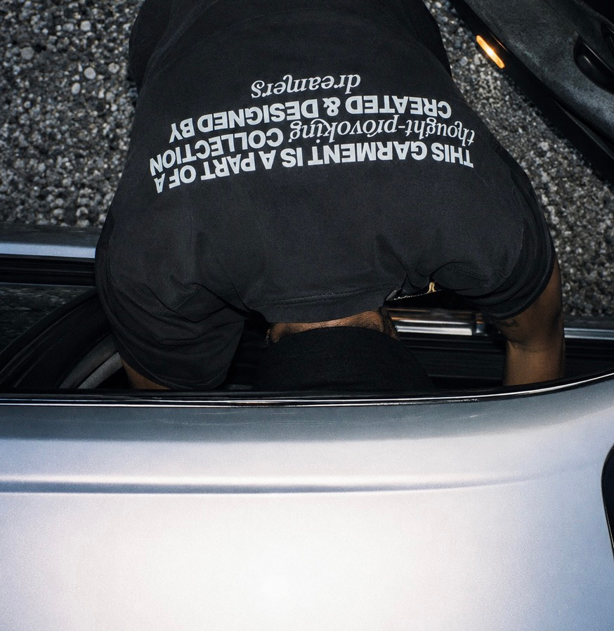 A person leaning into a car trunk, wearing a black t-shirt with white text on the back. The background shows gravel ground.