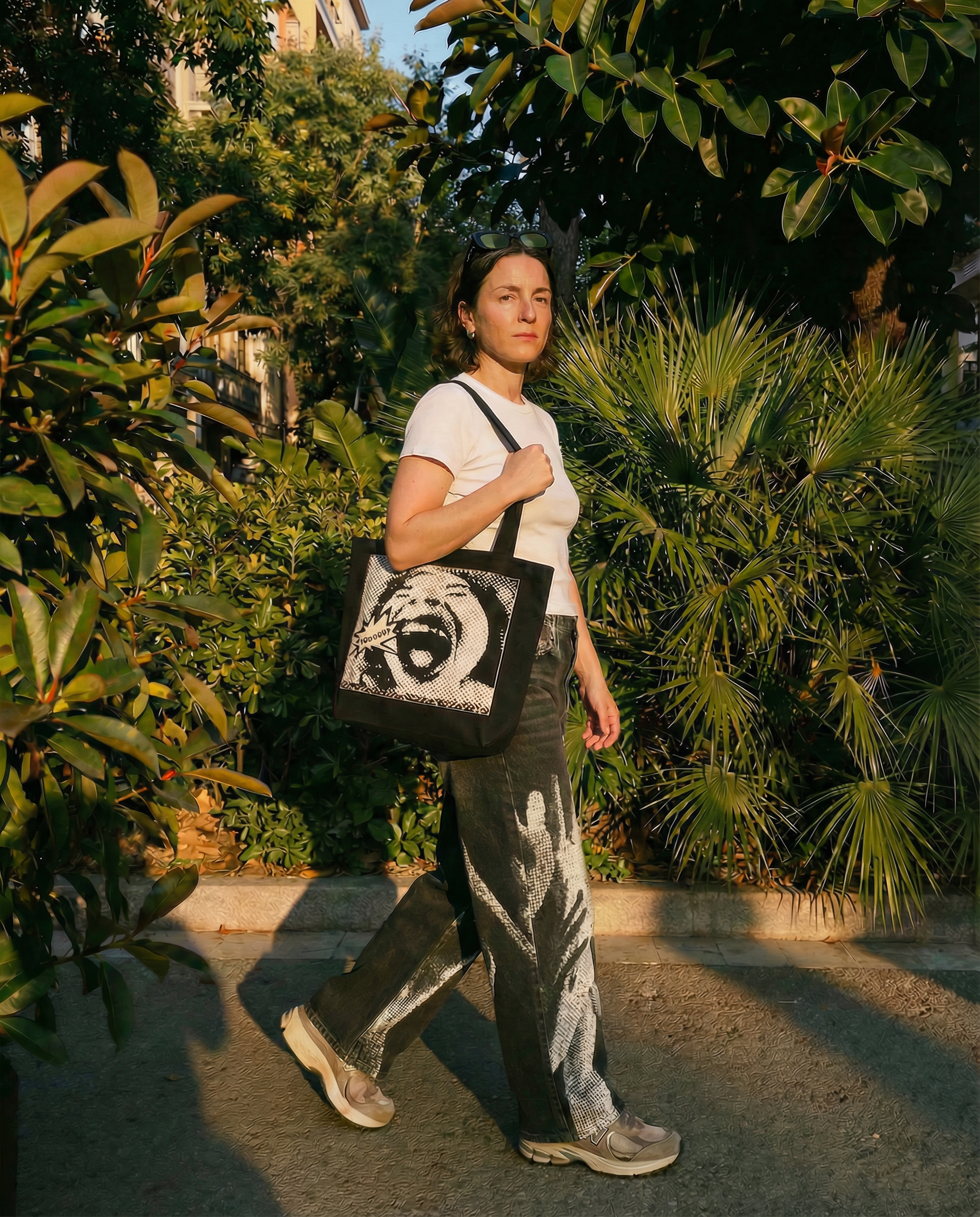 A woman standing outdoors among green bushes and trees, wearing a white T-shirt, patterned pants, sneakers, and sunglasses on her head, carrying a tote bag with a graphic of a laughing face and text.