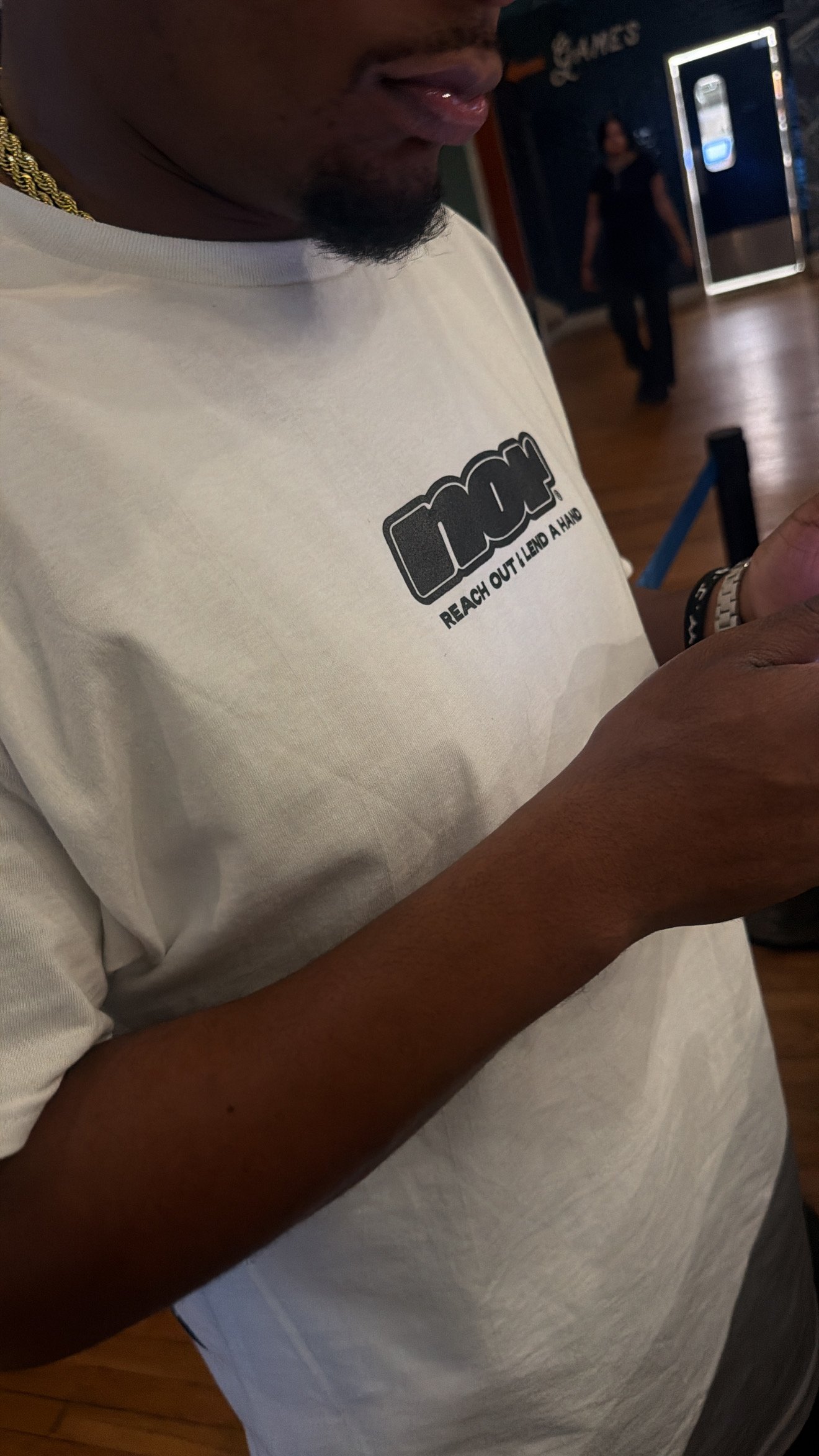 A person wearing a white T-shirt with a logo that says 'Reach Out I Lend A Hand' and gold chains, looking at a smartphone in a public indoor area.