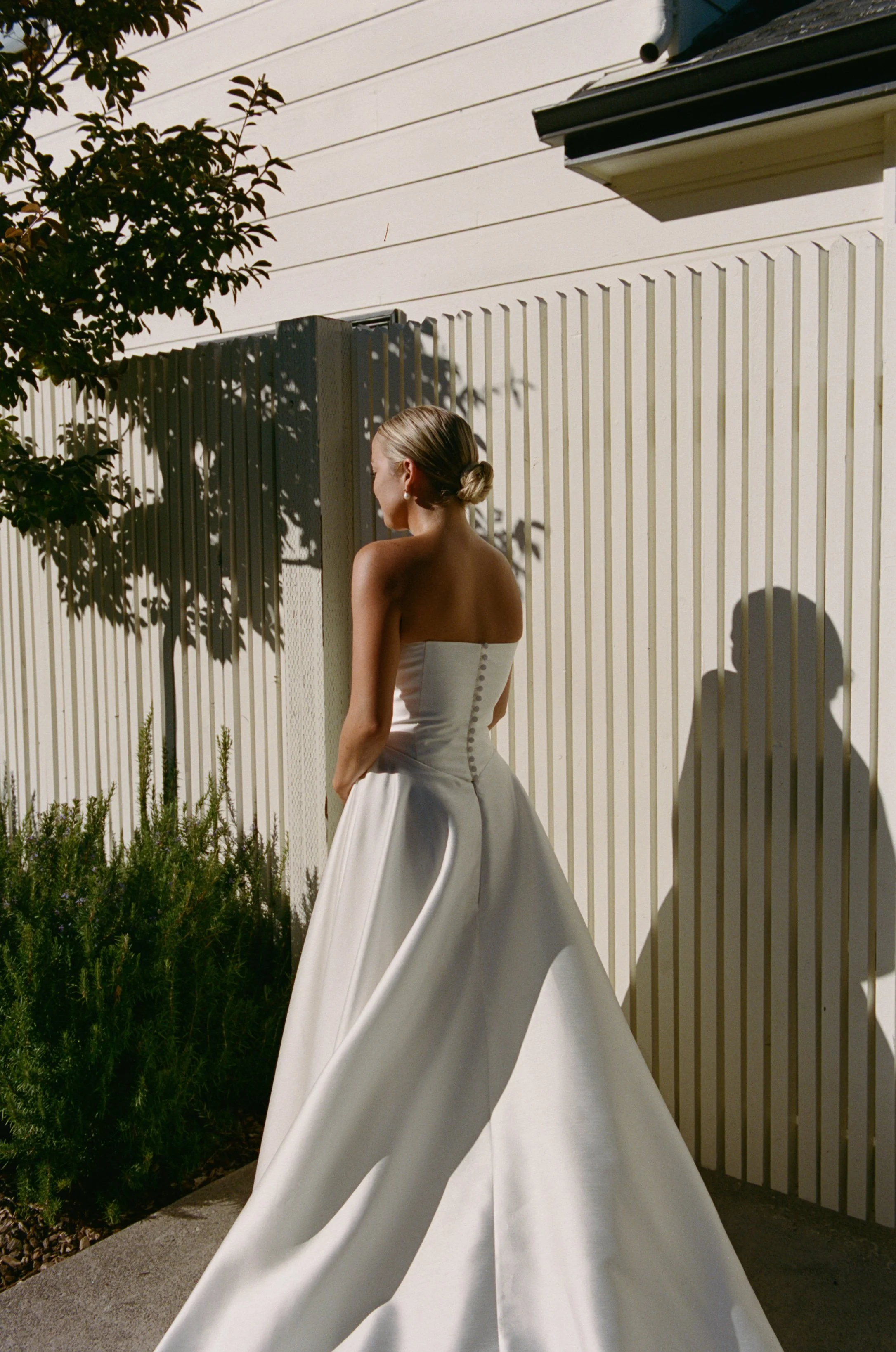 A woman in a white strapless wedding gown standing outdoors, with her back to the camera, near a fence and greenery, casting a shadow on the fence.