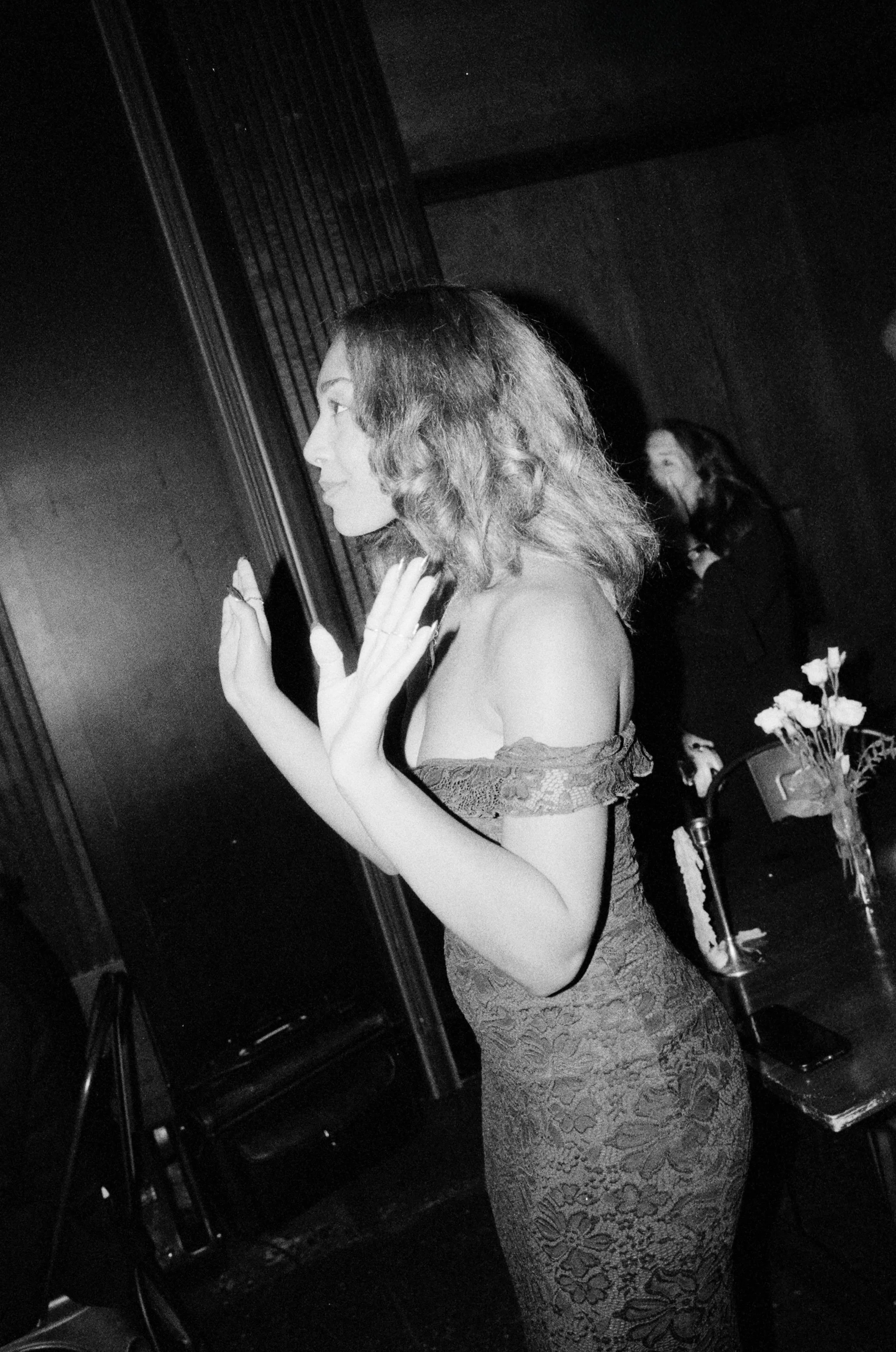A woman with shoulder-length wavy hair and wearing an off-the-shoulder lace dress gestures with her hands in a black and white photo, standing indoors near a dark wall and a small table with a flower arrangement and a smartphone.