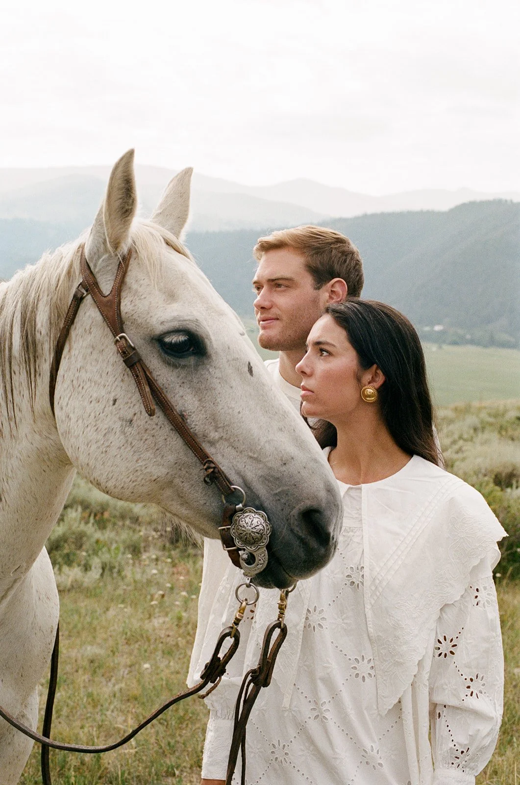 A man and a woman stand closely behind a white horse in an open field with mountains in the background. The man has short, light brown hair, and the woman has long, dark hair and gold earrings. Both are looking to the side.