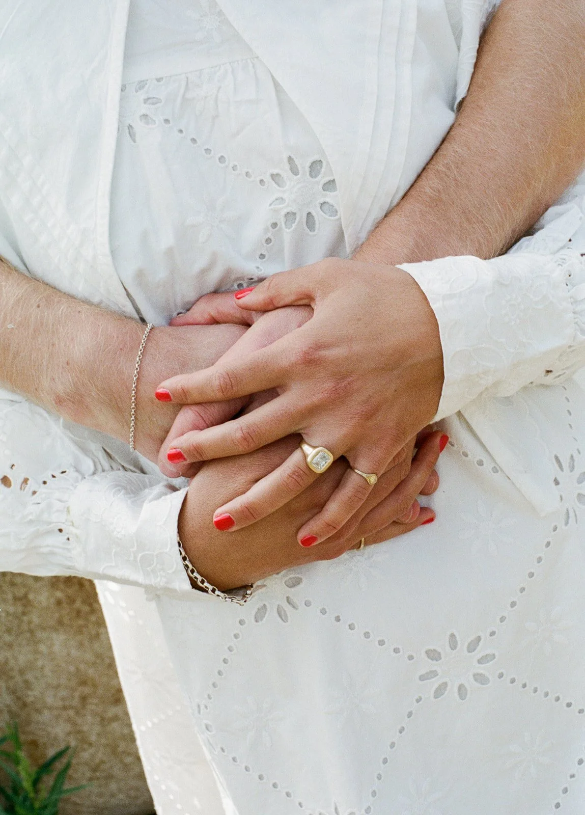 Multiple hands, gently holding each other, with the woman wearing jewelry and a white embroidered dress.
