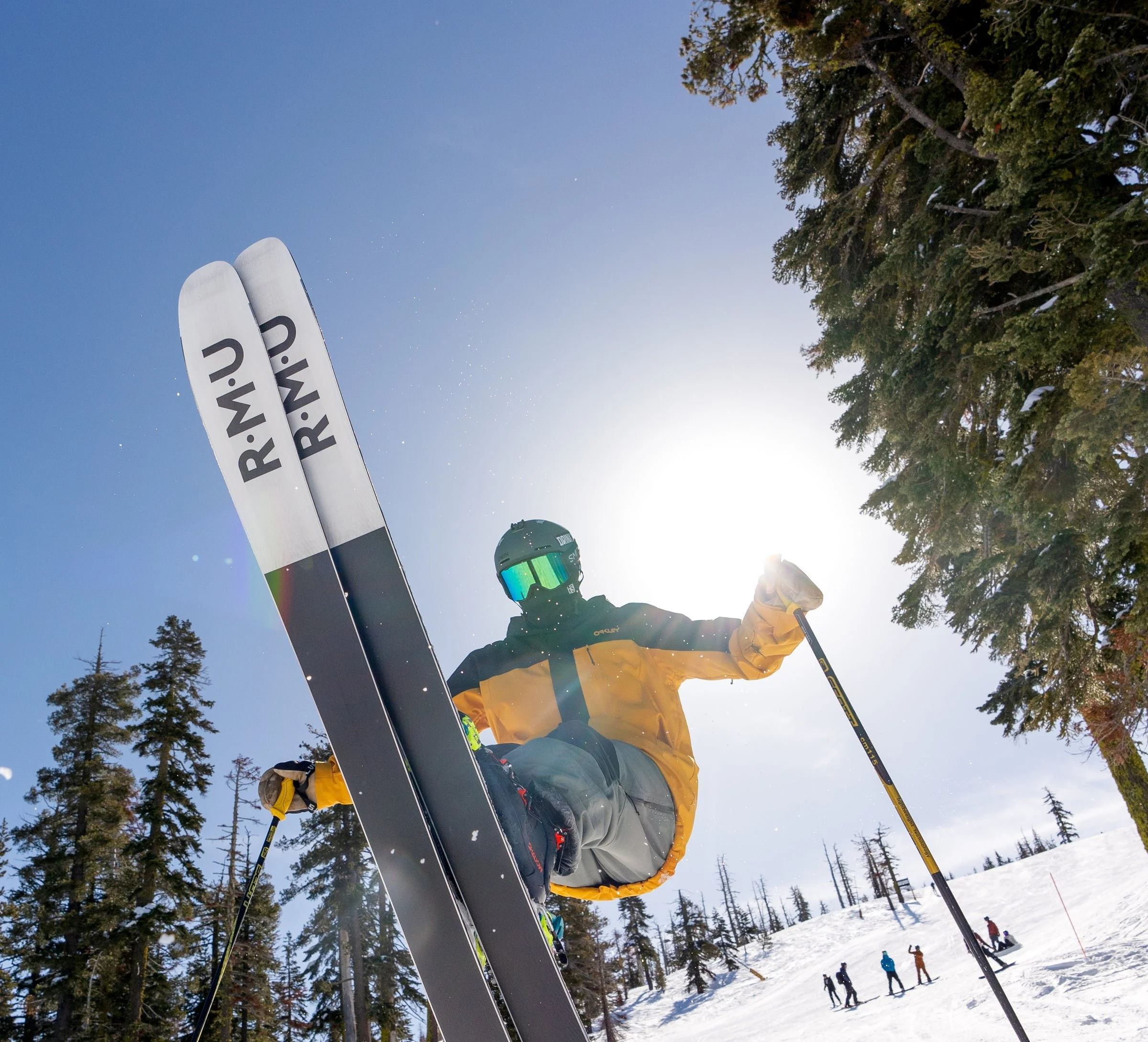A skier in a yellow and black jacket, gray pants, and a helmet with goggles, standing on snow with skis and poles, surrounded by trees and other skiers in the background under a clear blue sky.