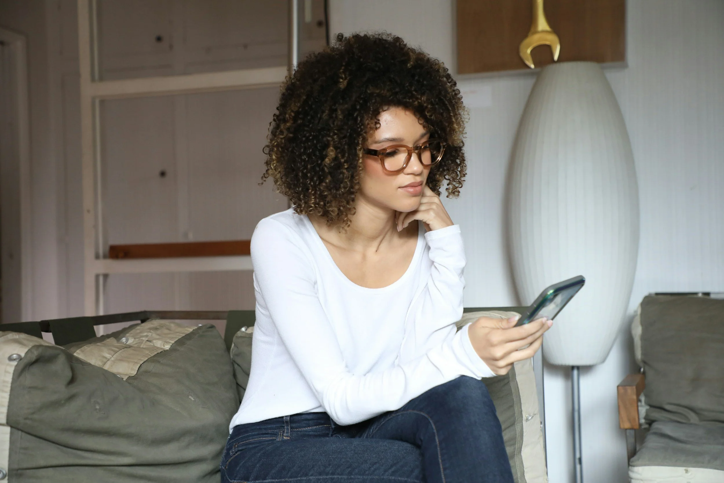 Woman with curly hair sitting on a sofa, looking at her phone with a thoughtful expression, wearing glasses and a white top.