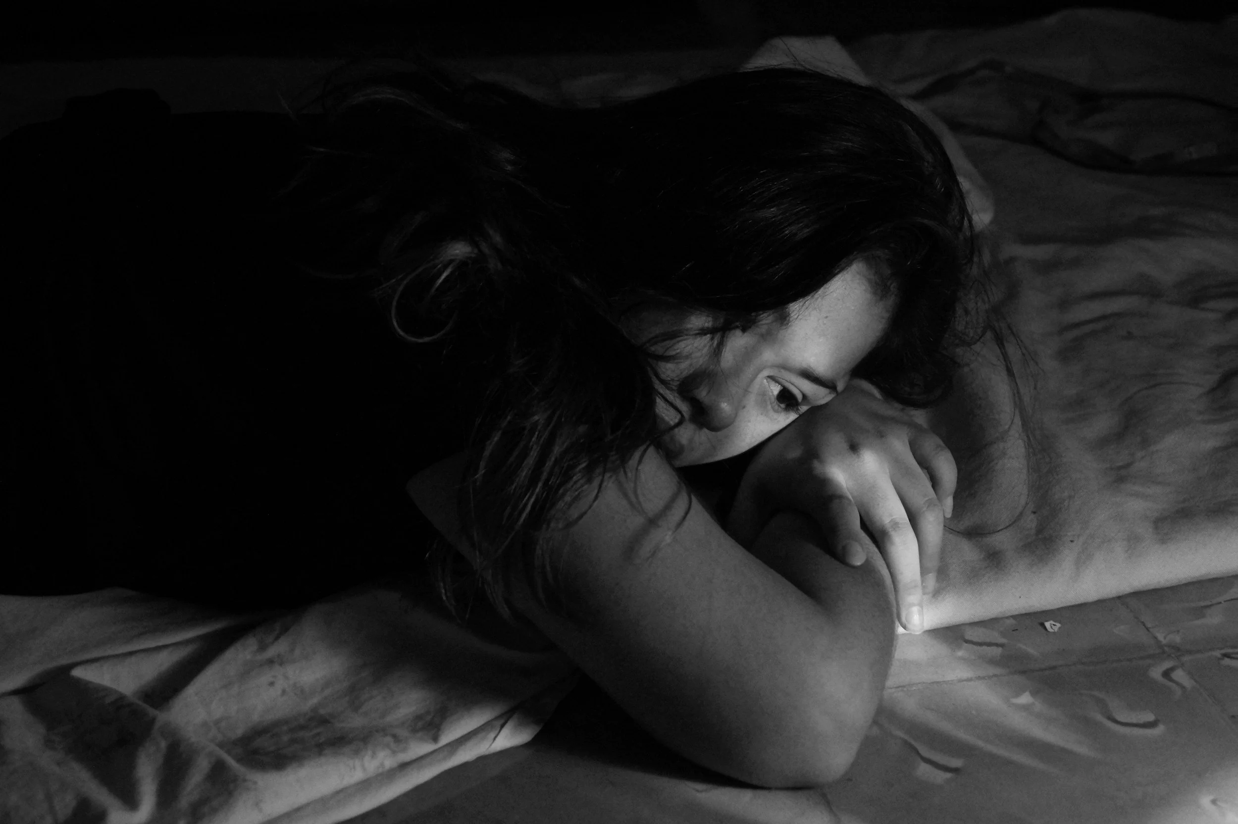 Woman lying curled up in bed in low light, representing the emotional impact of trauma, nervous system overwhelm, and the long-term effects of chronic stress.