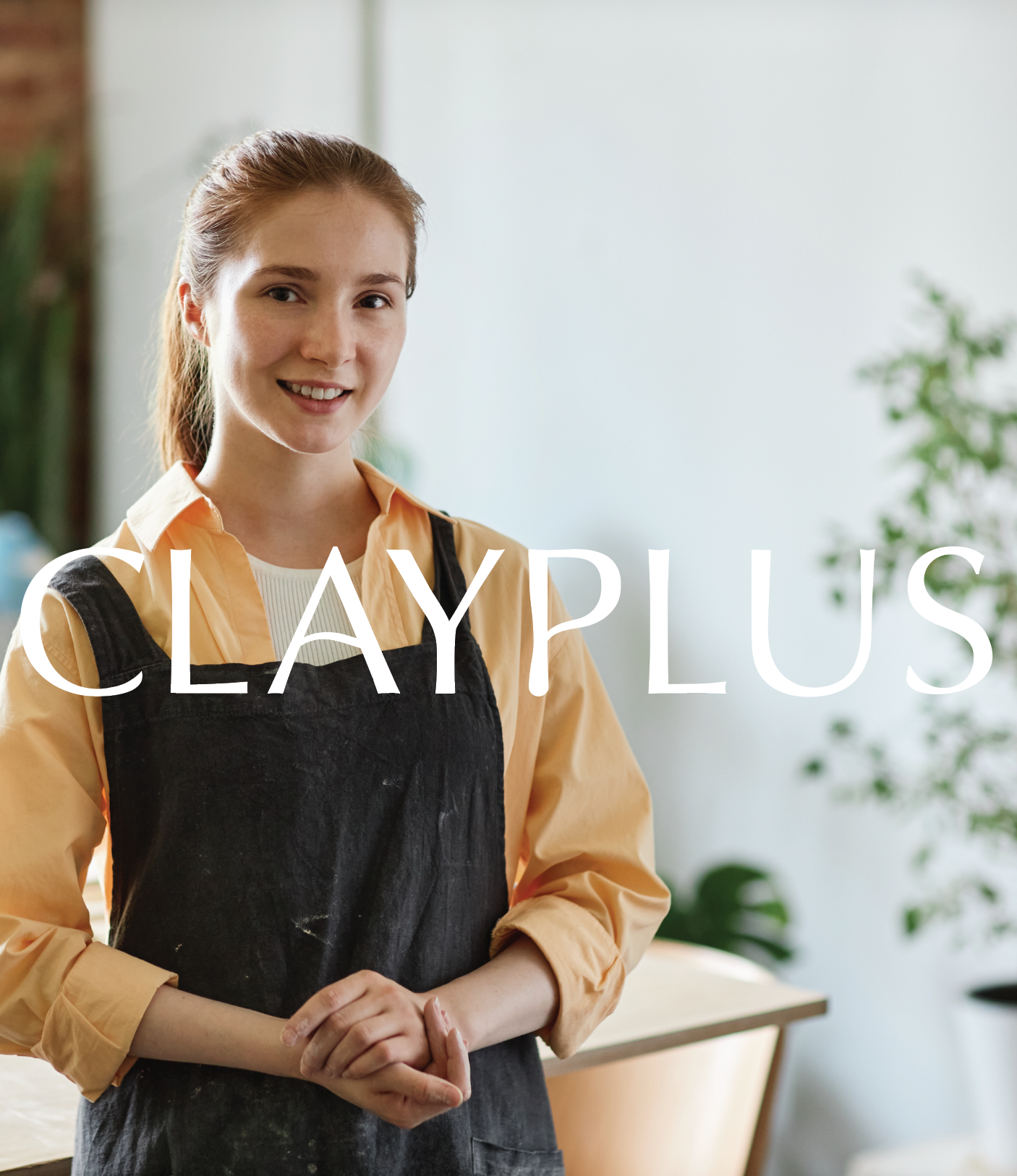 A young woman with red hair wearing a beige shirt and black apron, smiling indoors with plants in the background.