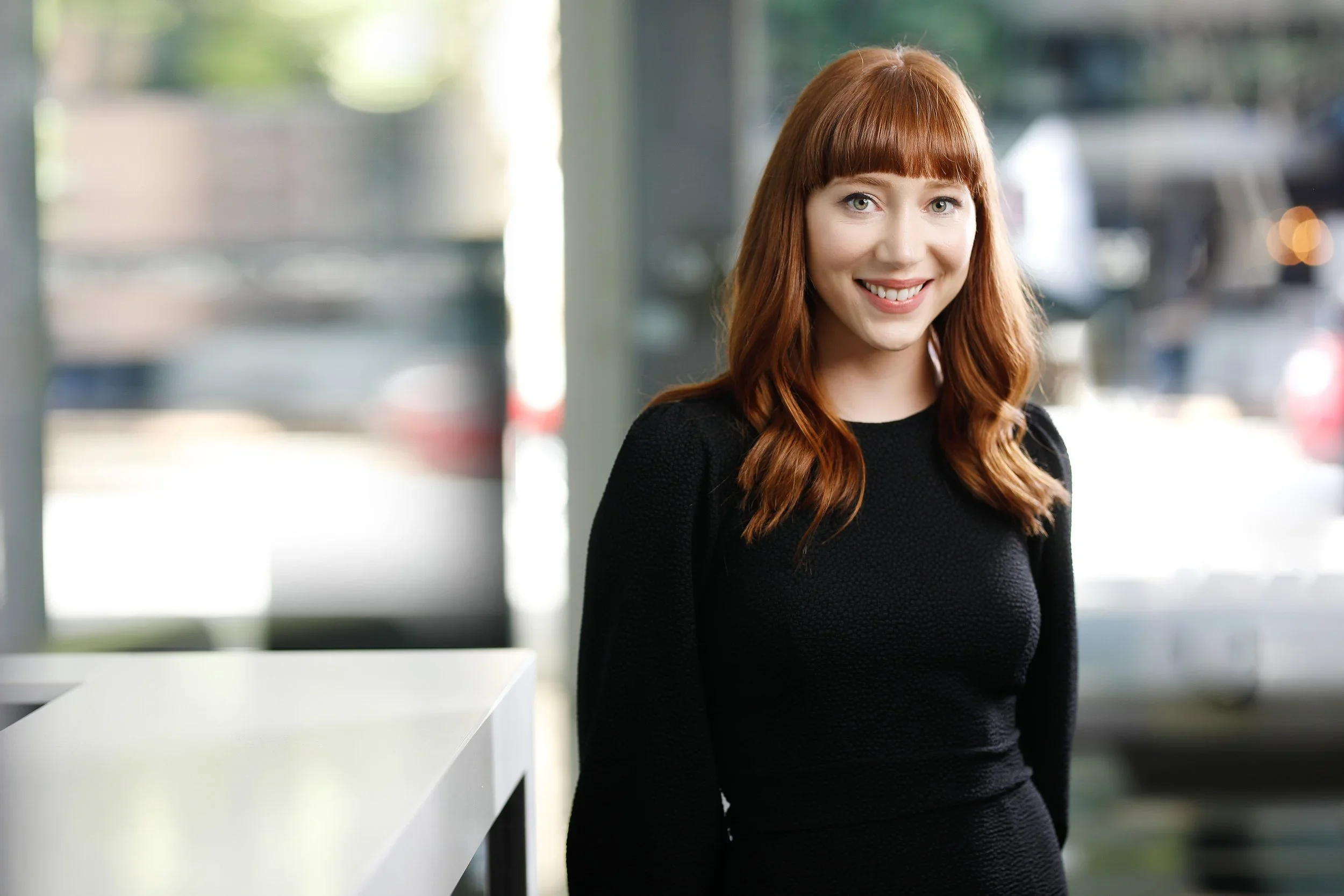 A young woman with red hair and green eyes smiling, wearing a black top, standing indoors near a large window with blurred outside scenery.