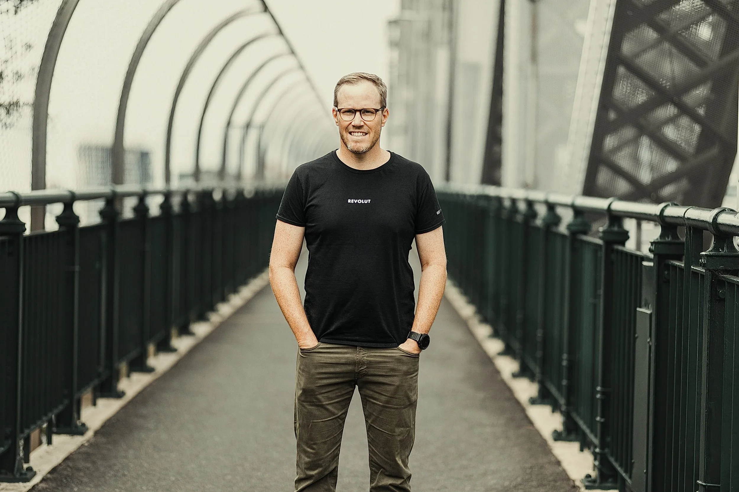 A smiling man with glasses and a black T-shirt standing on a bridge or walkway with railings, with a metal arch structure overhead.