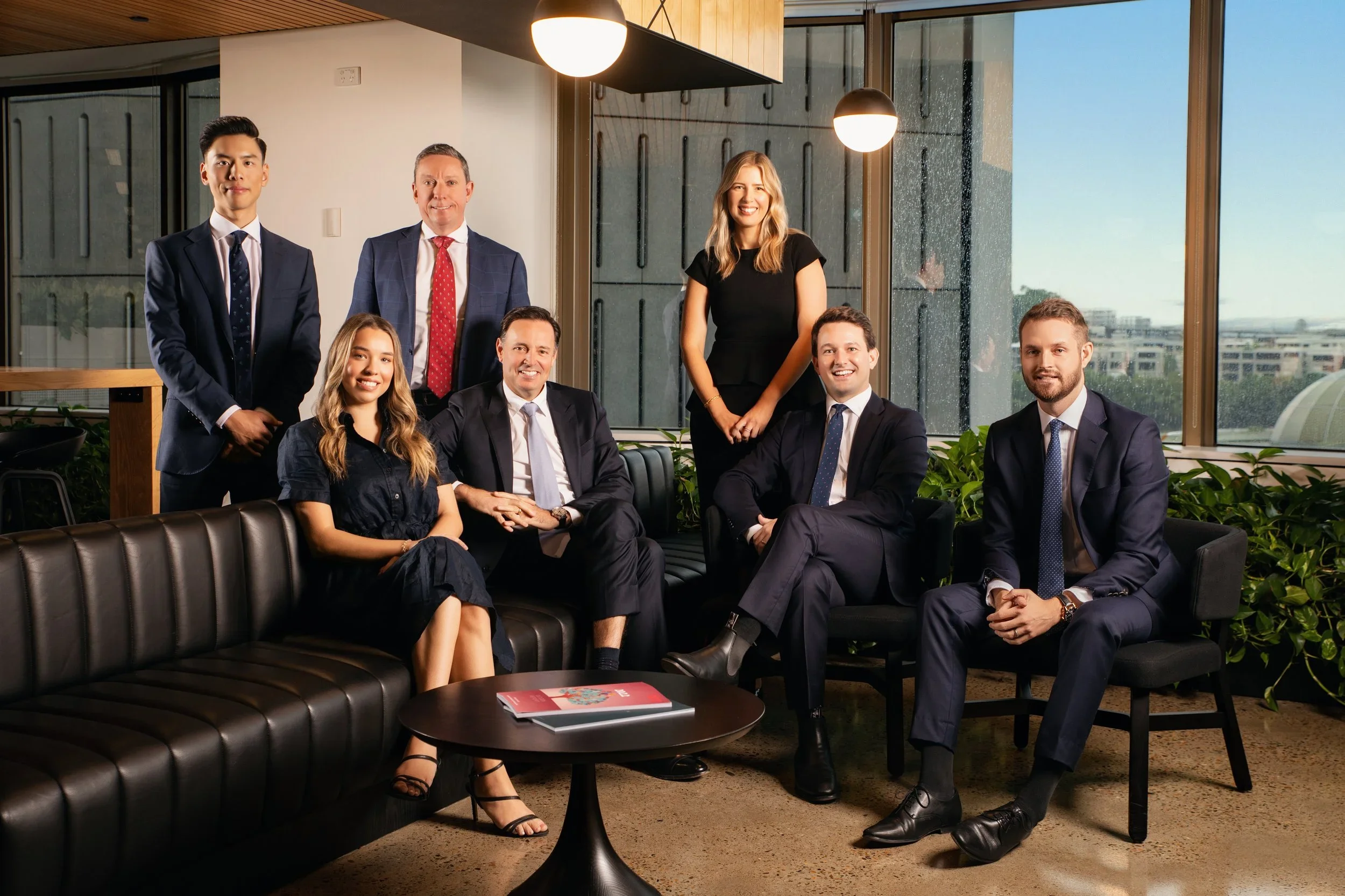A diverse group of eight professionals, four men and four women, posed in a modern office with large windows, indoor plants, and pendant lighting, smiling for the camera.