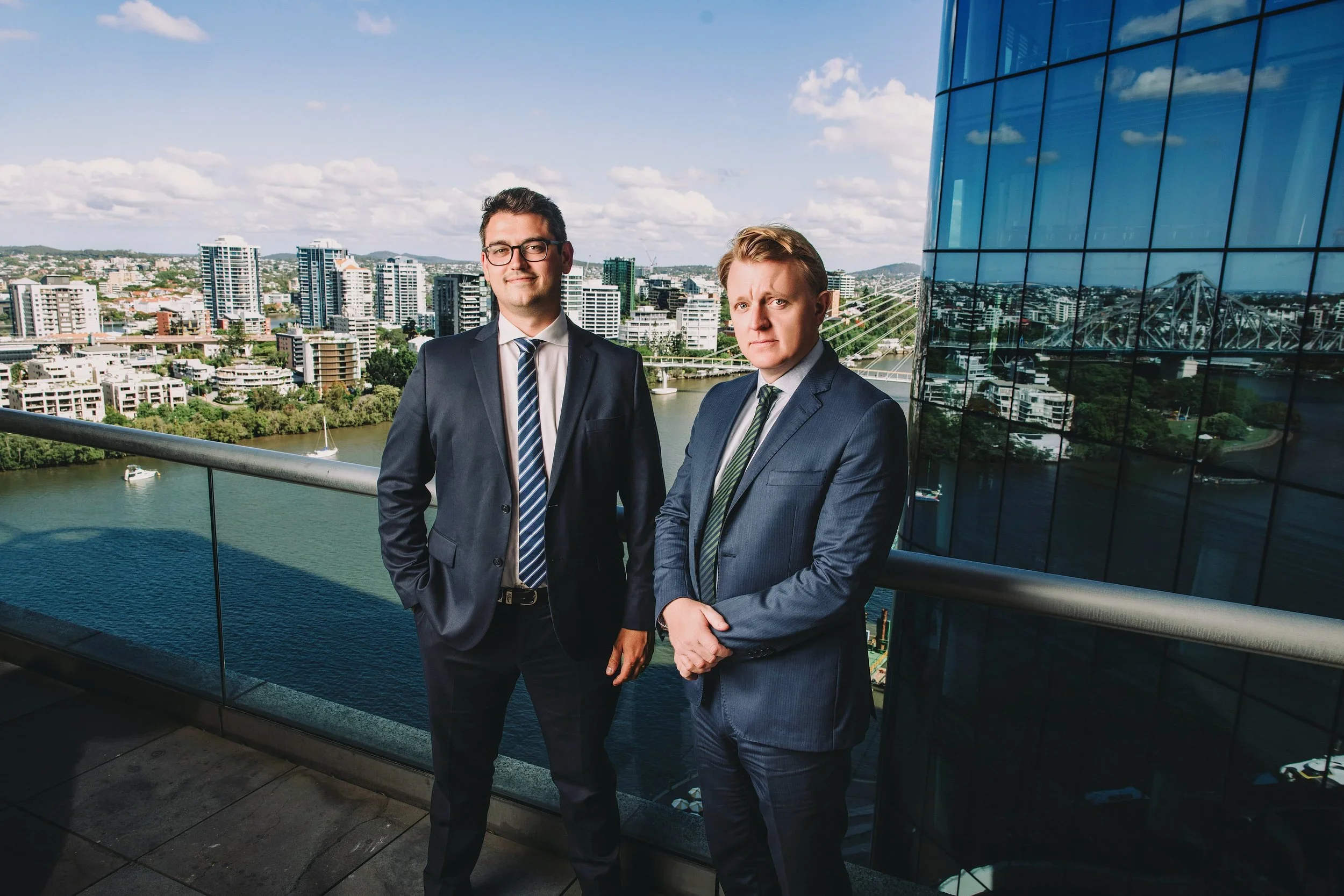 Two men in suits standing on a balcony overlooking a cityscape with modern buildings, a river, and a blue sky with clouds.