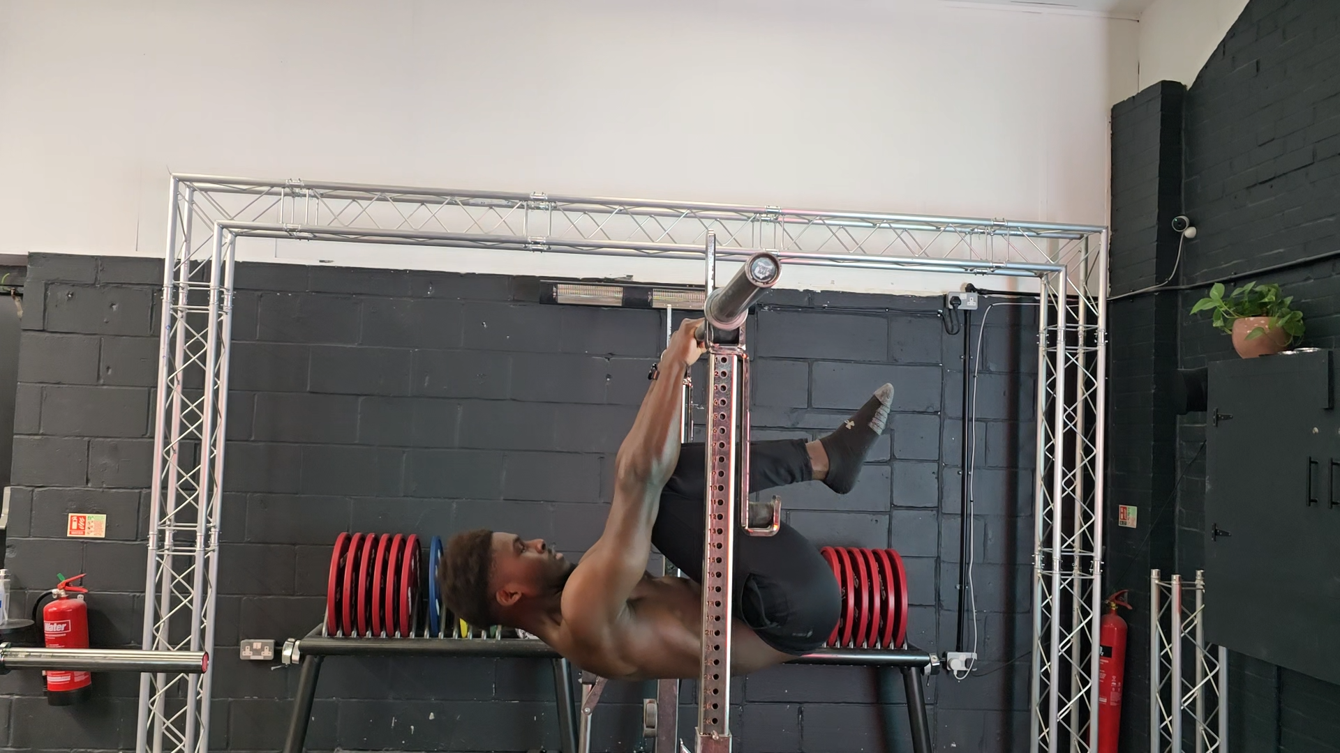 A shirtless man in black shorts lying on a bench, holding a barbell overhead with one leg raised in a gym with black walls and metal scaffolding.