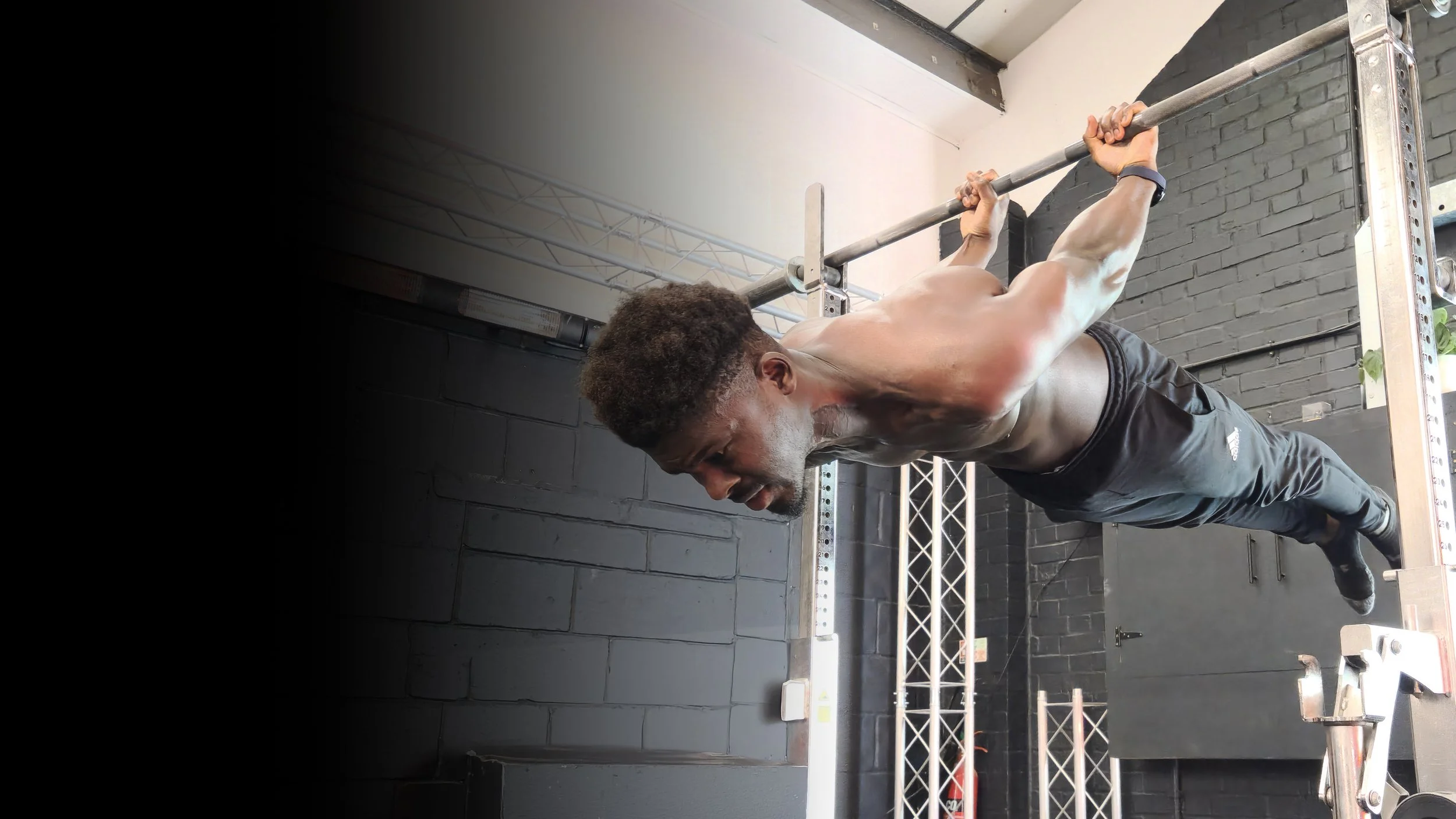 A shirtless man with dark hair and a beard performing a pull-up exercise on a metal bar, in a gym with black brick walls and workout equipment.