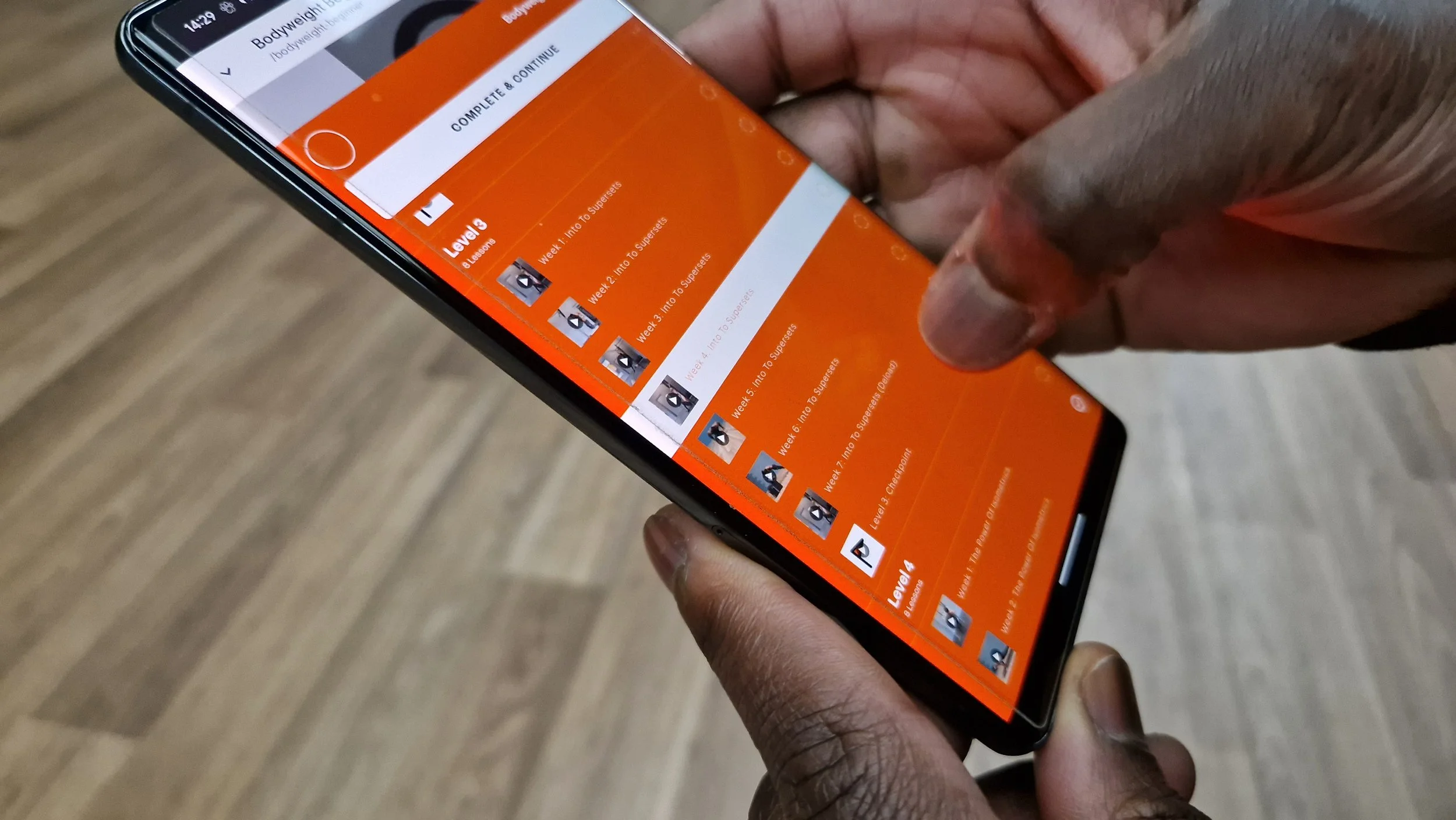 A person holding a smartphone displaying a workout app with a bright orange interface showing exercise levels and weekly progress.