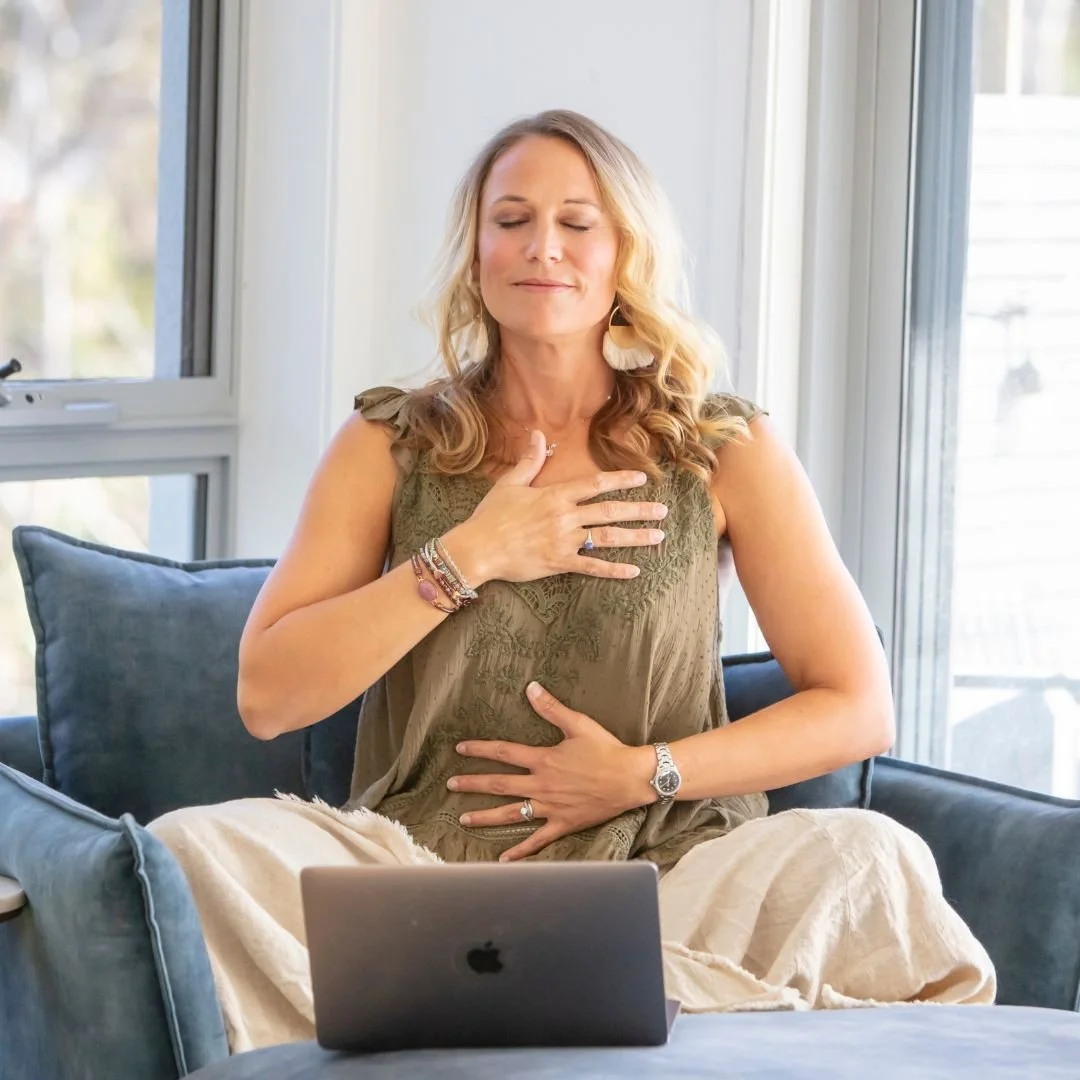 A woman with blonde hair sitting on a couch with her eyes closed, touching her chest and stomach, in front of a laptop inside a bright room with windows.