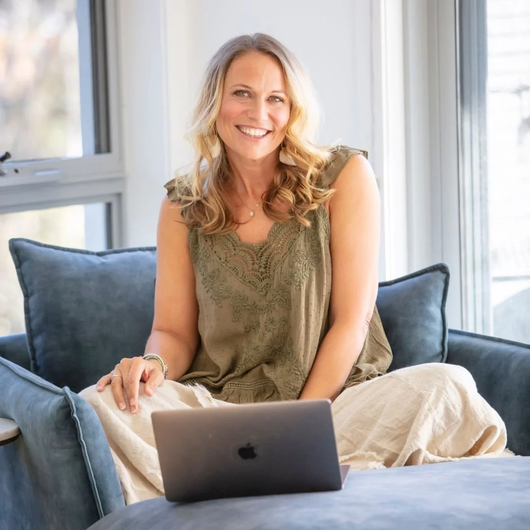 A woman with blonde hair smiling while sitting on a couch with a laptop in her lap, in a bright room with large windows.