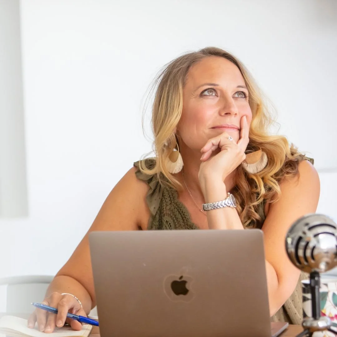 A woman sitting at a desk with a laptop and microphone, looking thoughtful and smiling.