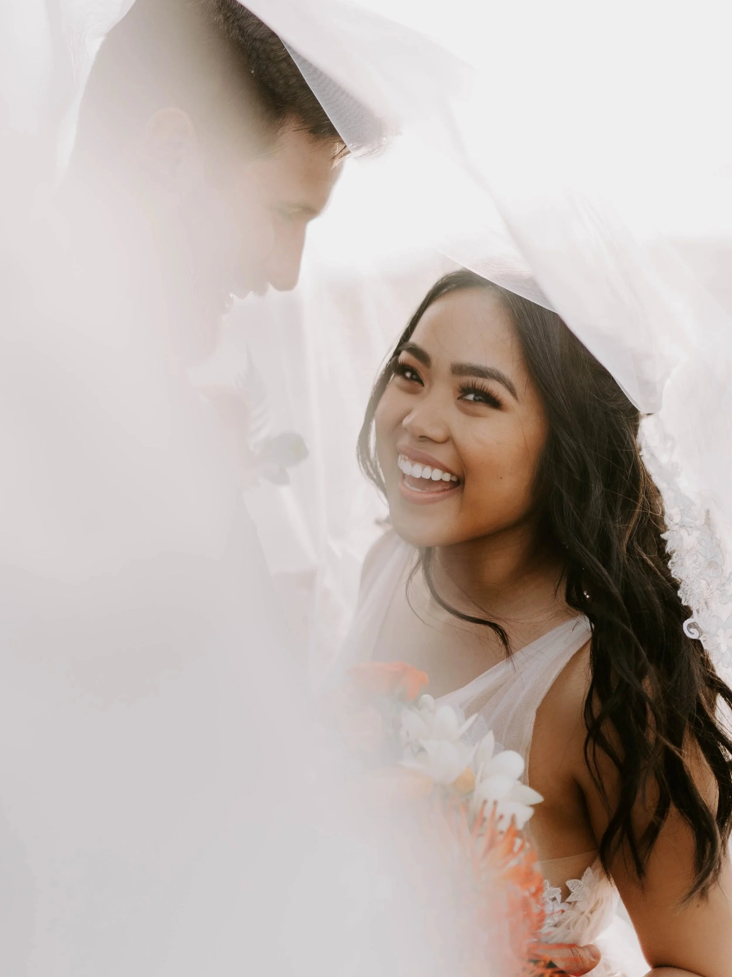 A smiling bride and groom on their wedding day, with the bride wearing a wedding dress and veil, and the groom in a tuxedo, under a piece of white fabric or veil.