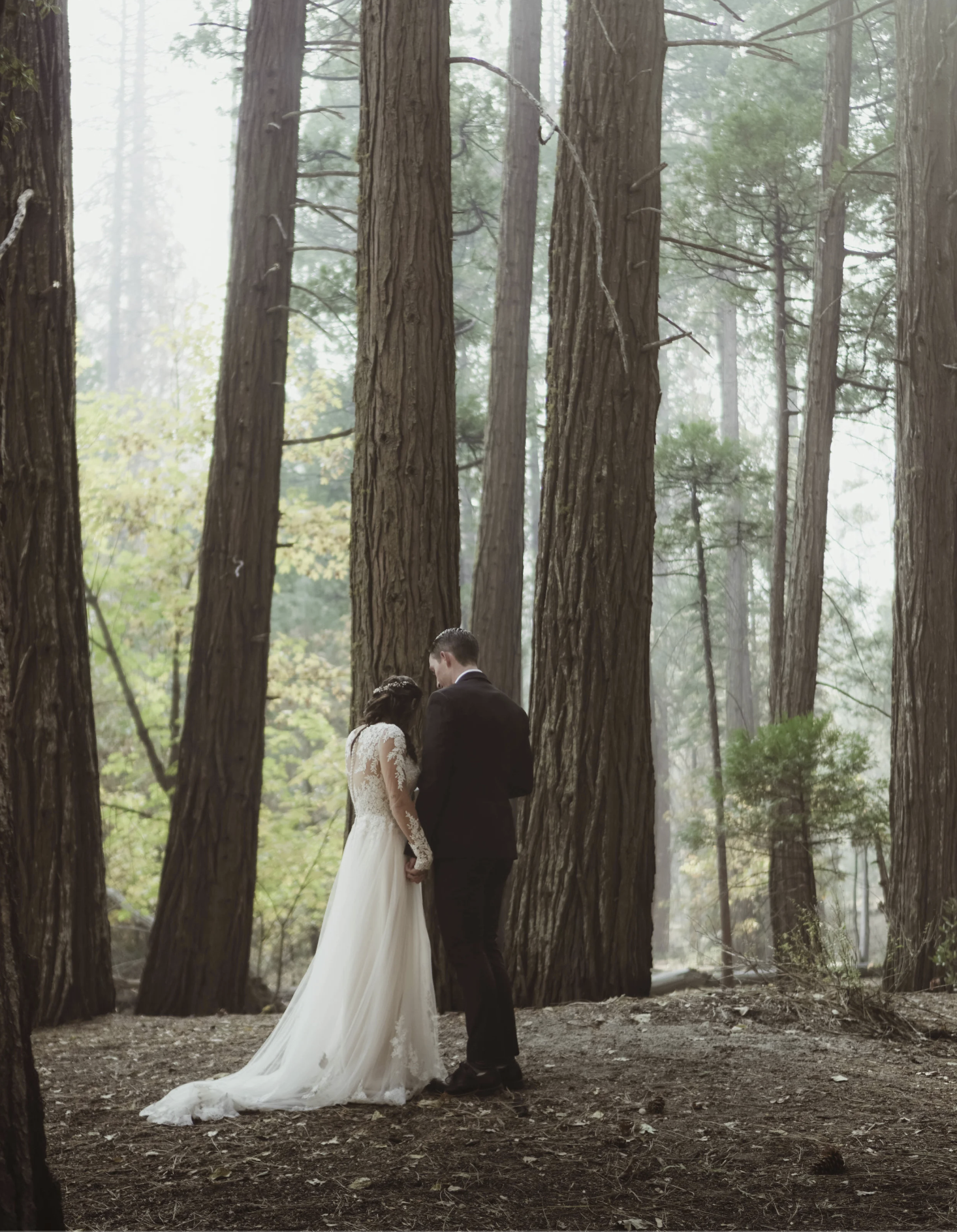 A bride and groom standing closely together in a forest during a wedding ceremony, holding hands while praying with their head bowed.