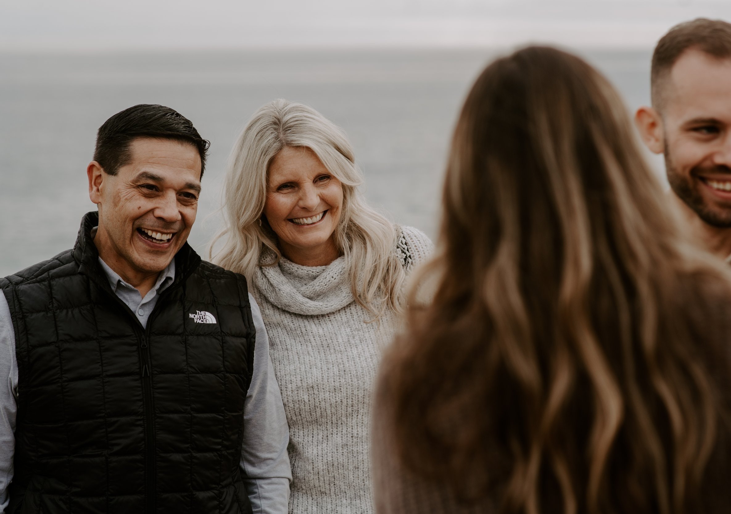Group of people smiling outdoors near water, with two women and two men, one woman with blonde hair and a man in a black vest.