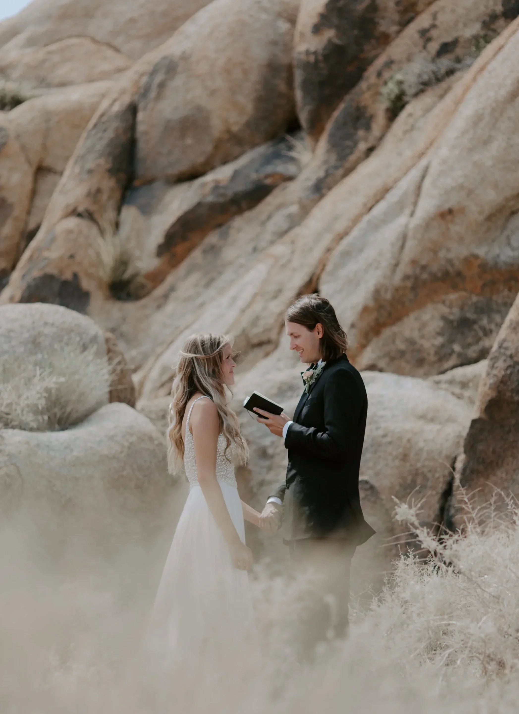 A couple in wedding attire stands holding hands in a desert landscape with large rocks, as the groom reads vows from a small black book.