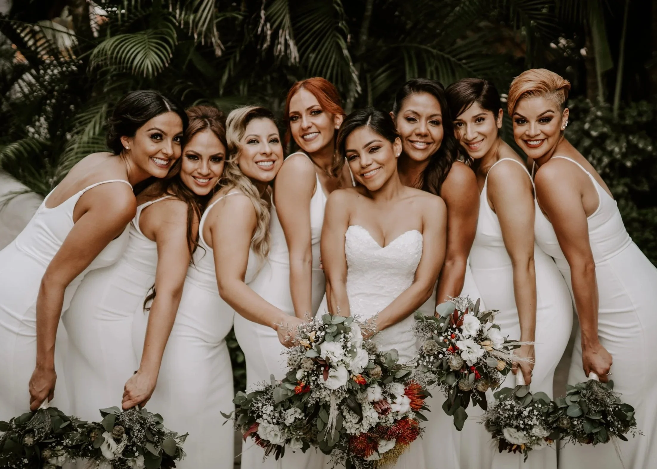 A group of nine women in white dress, posing together outdoors with green foliage in the background, holding bouquets of flowers.