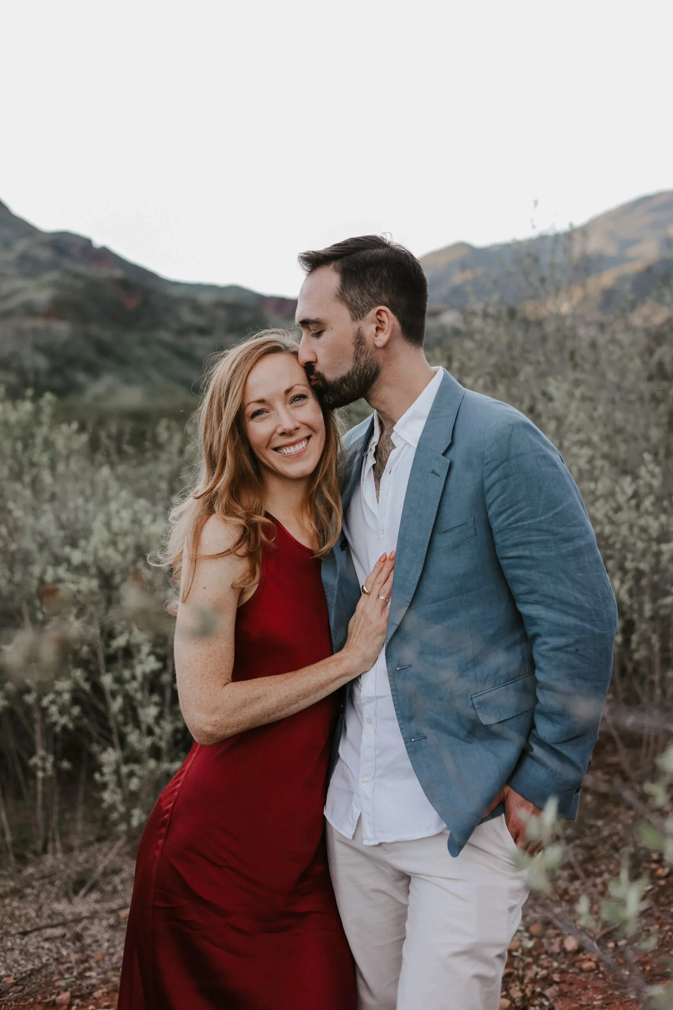 A smiling woman with long red hair wearing a red dress standing next to a man with dark hair, beard, wearing a blue blazer and white shirt, sharing a kiss on the forehead outdoors with mountains and bushes in the background.