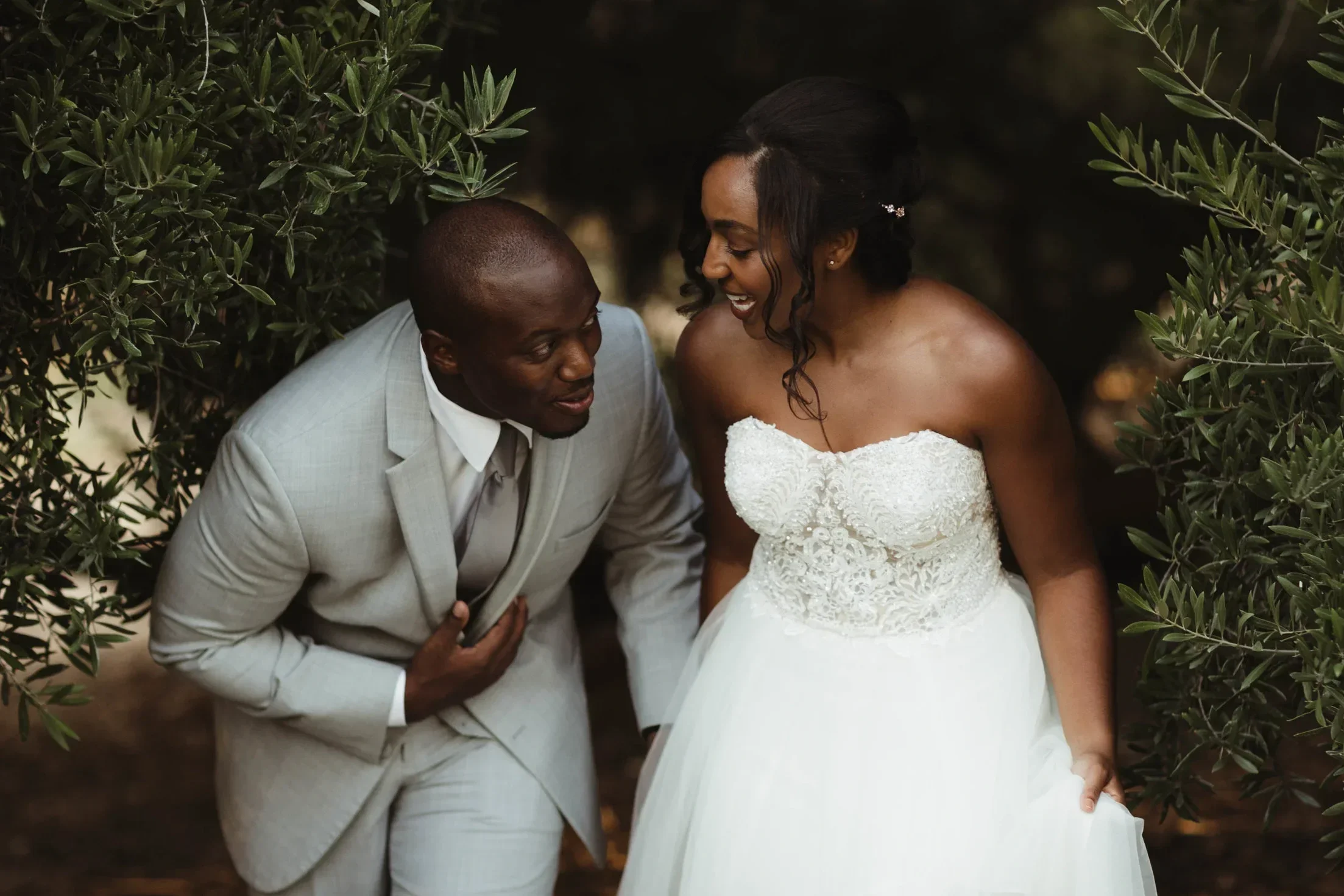 A bride and groom laughing and enjoying each other's company outdoors amidst greenery.