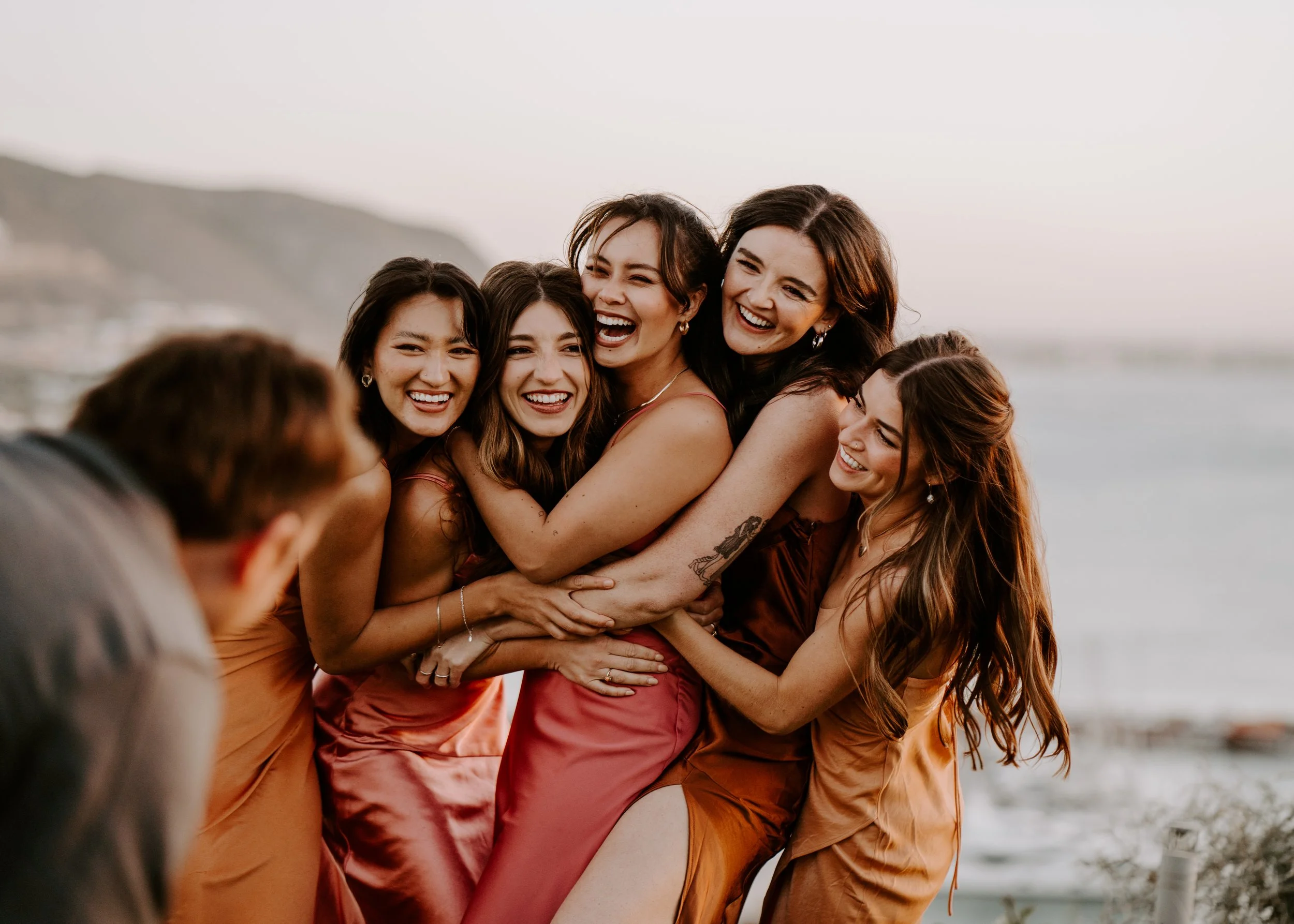 Group of women wearing satin dresses, hugging and laughing on the beach at sunset.