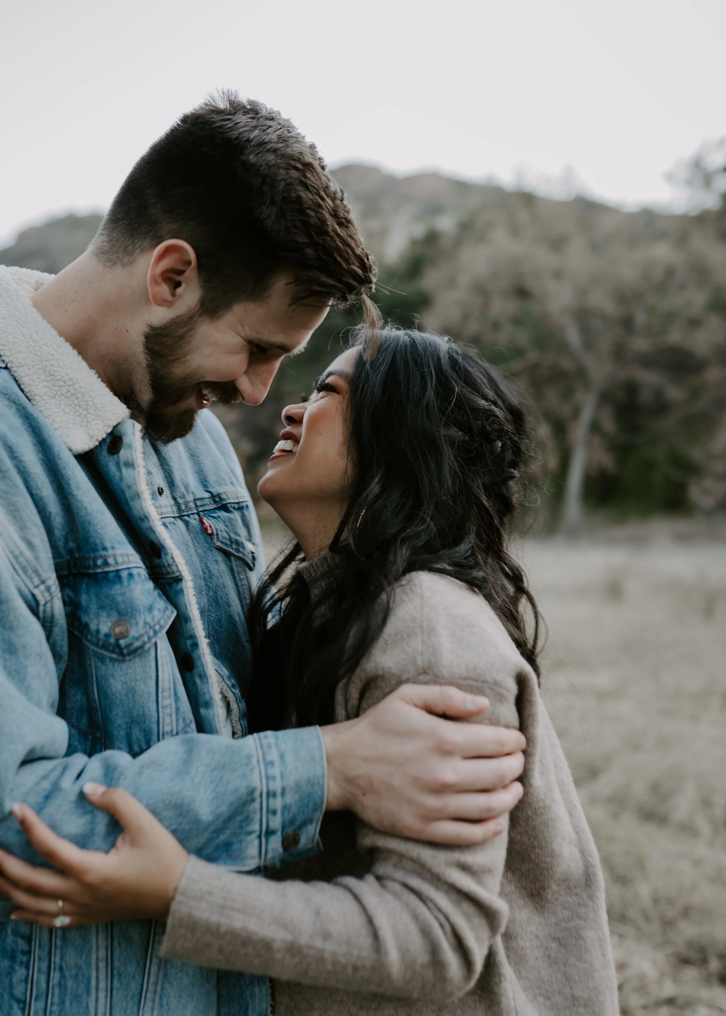A happy couple outdoors, smiling and looking into each other's eyes, embracing with mountains and trees in the background.