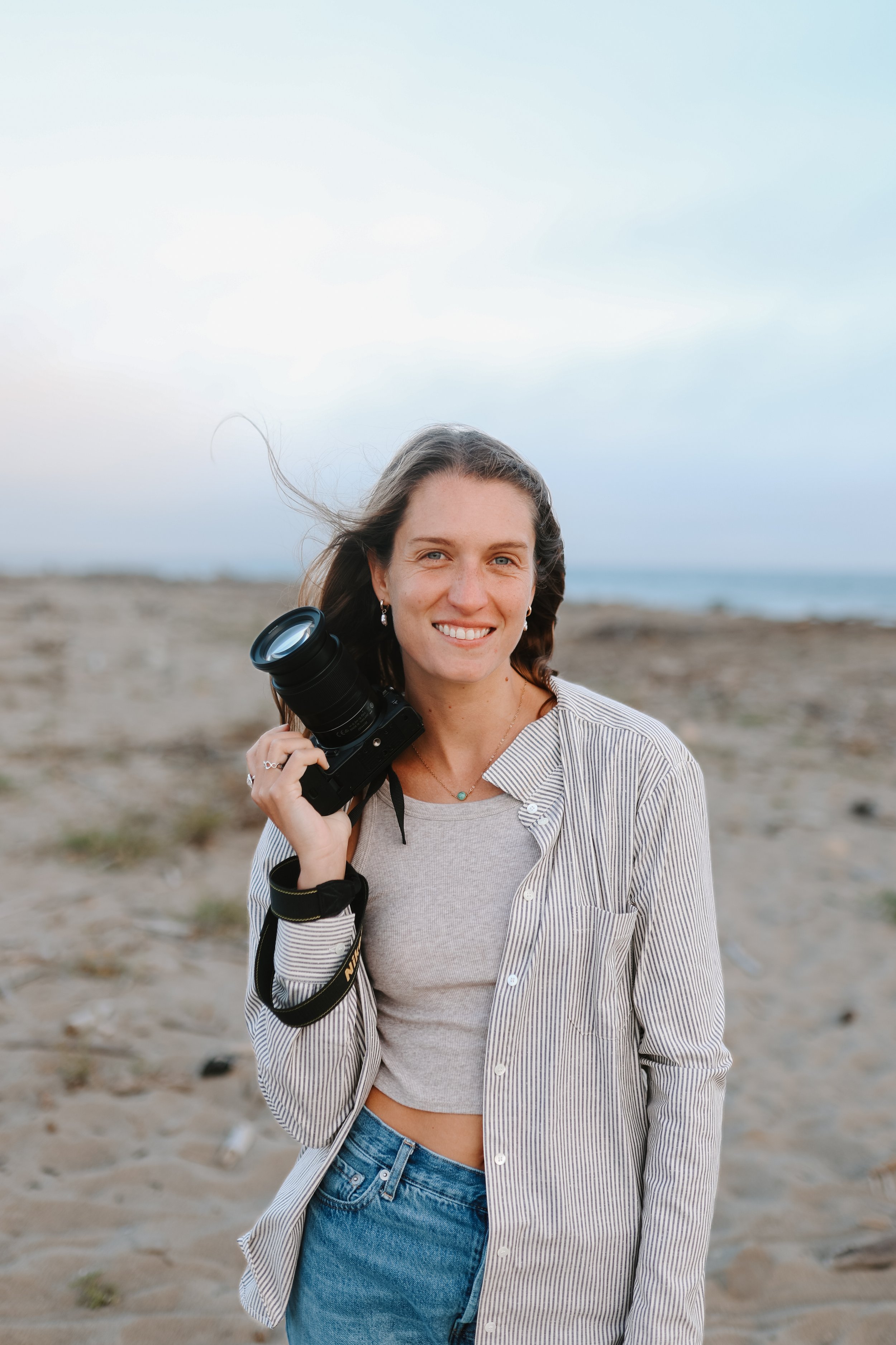 A young woman with long brown hair smiling and holding a camera on her shoulder at the beach.