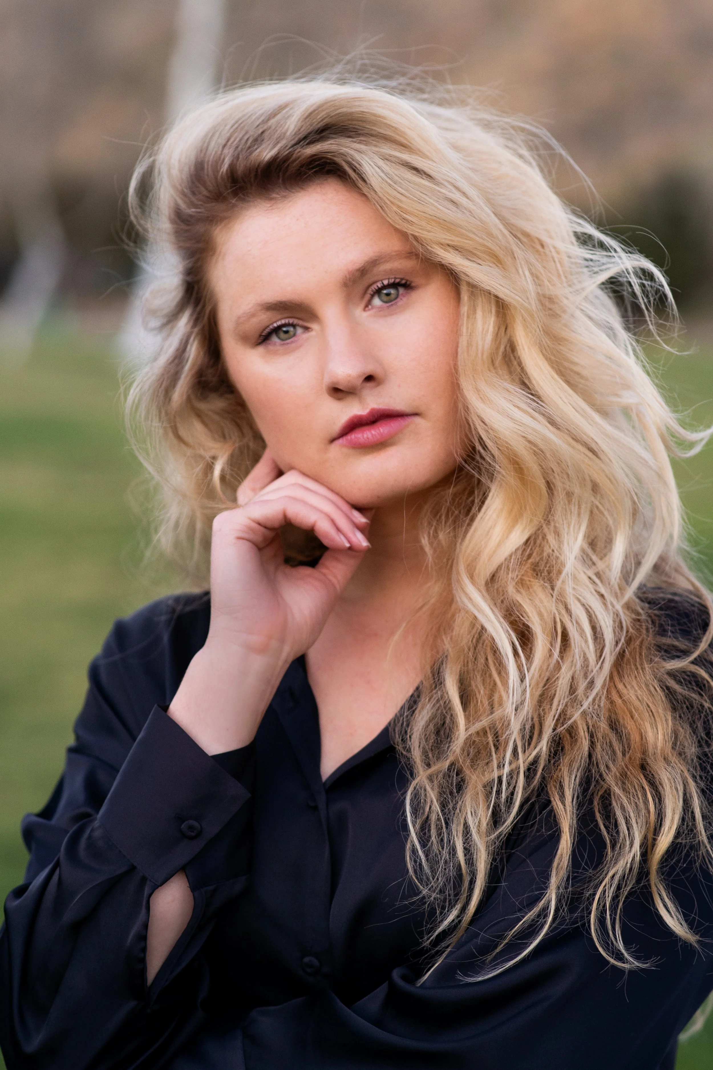 A portrait of a young woman with long, wavy blonde hair, wearing a black shirt, outdoors with blurred greenery in the background.